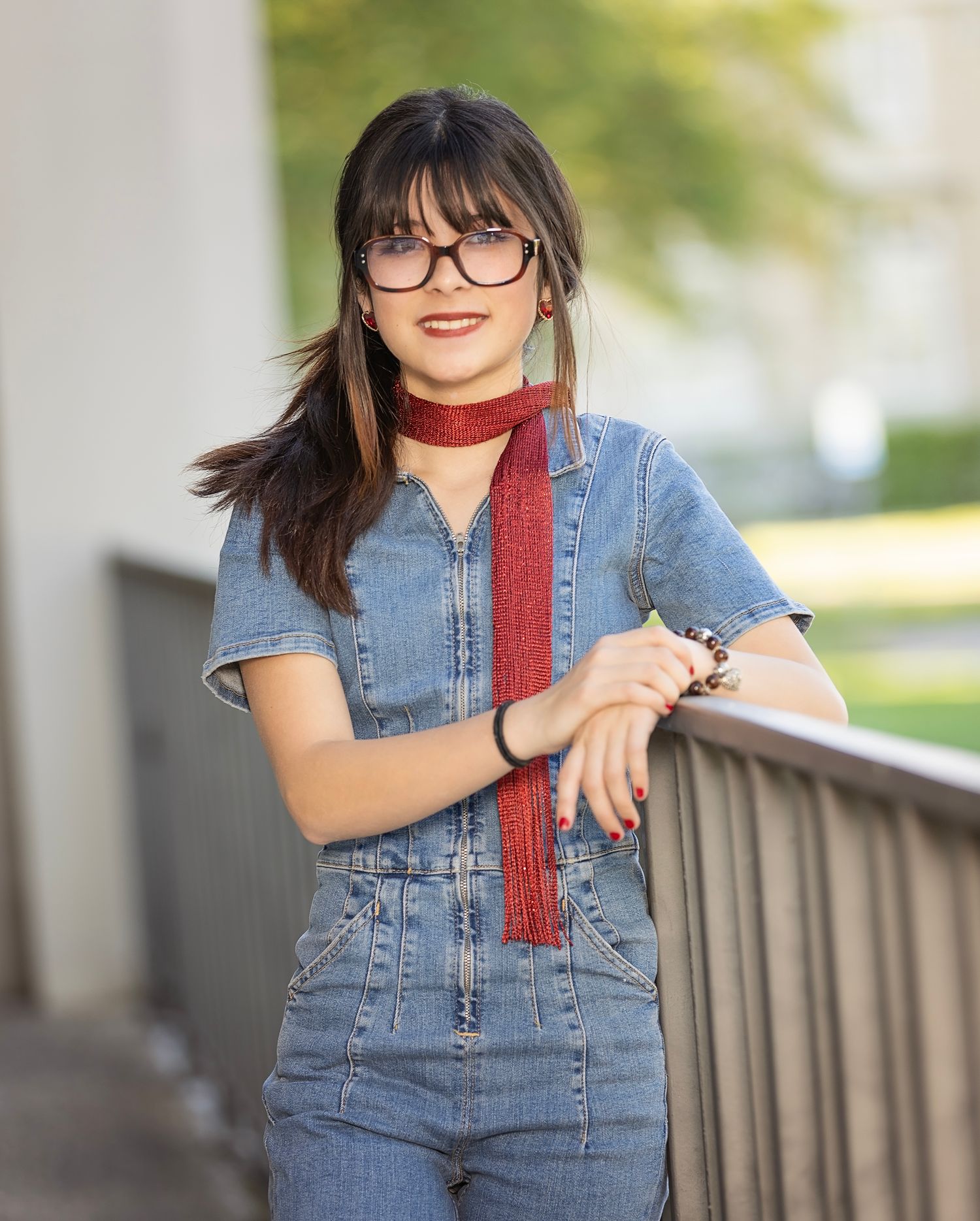 High School Senior wearing a denim dress and red scarf poses casually against a railing in an outdoor setting.