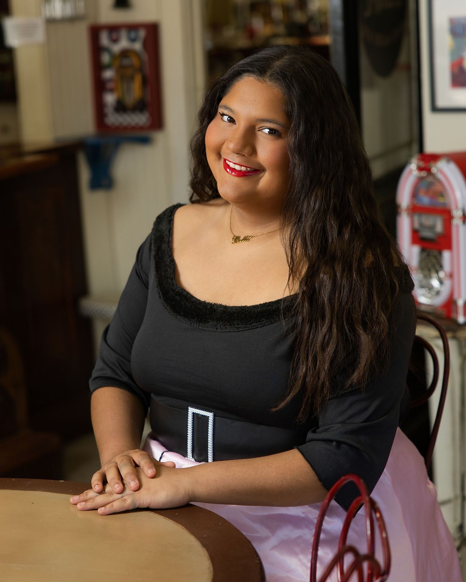 A person in a black dress sits at a table with a warm smile in an indoor setting.