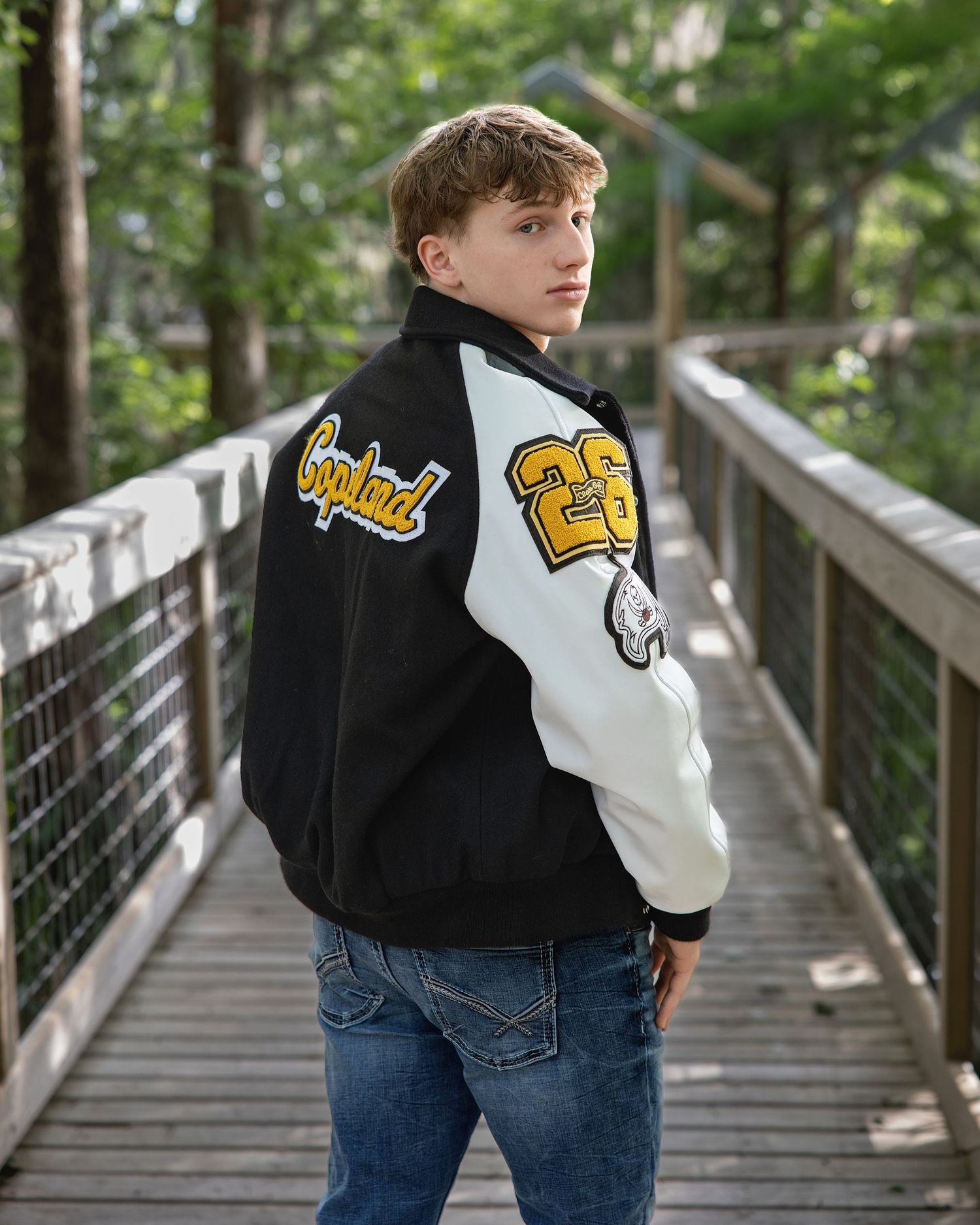 Teen boy in black and white varsity jacket with number 23 patch poses on wooden bridge surrounded by trees.