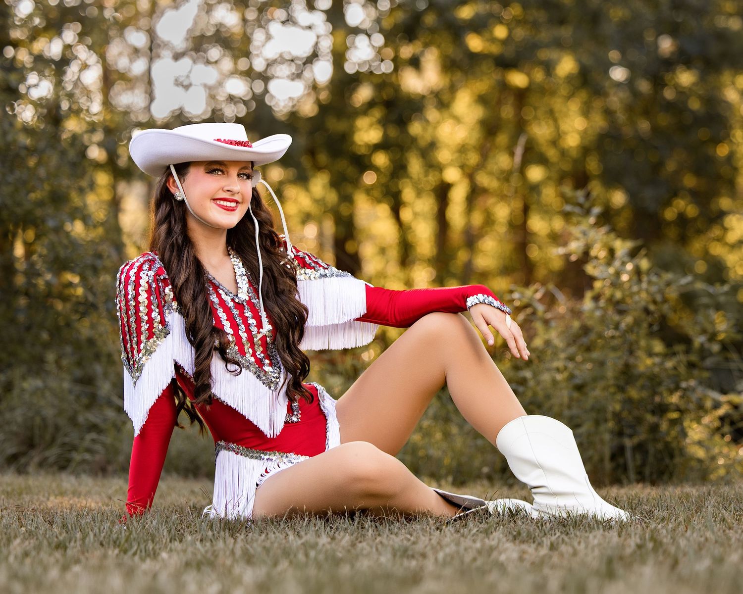 Young woman in red and white cowgirl outfit with white boots and hat sitting on grass outdoors.