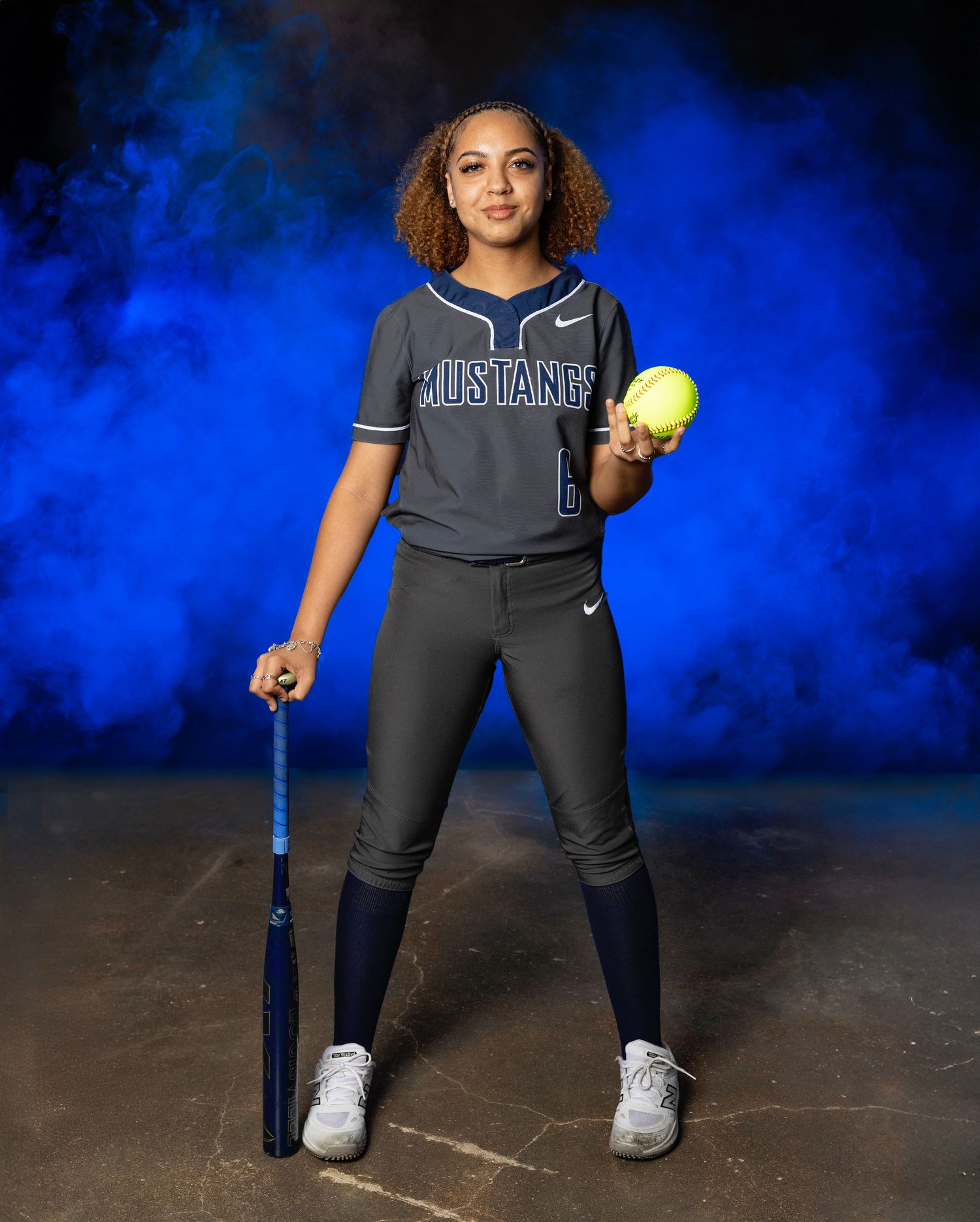 A softball player in a dark uniform stands in a ready position with a yellow ball and bat against a blue background.