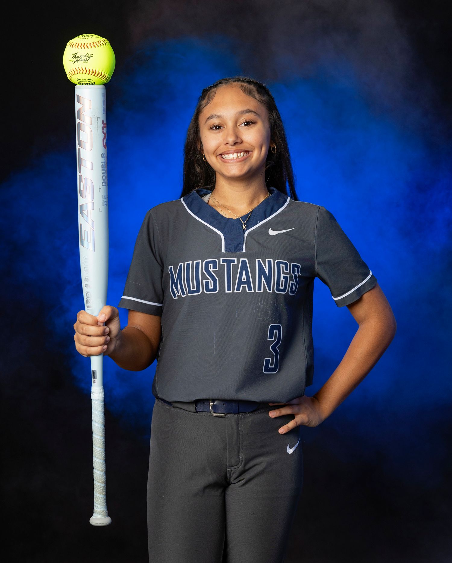 A softball player in dark gray uniform poses against a blue backdrop with sports equipment for team photos.
