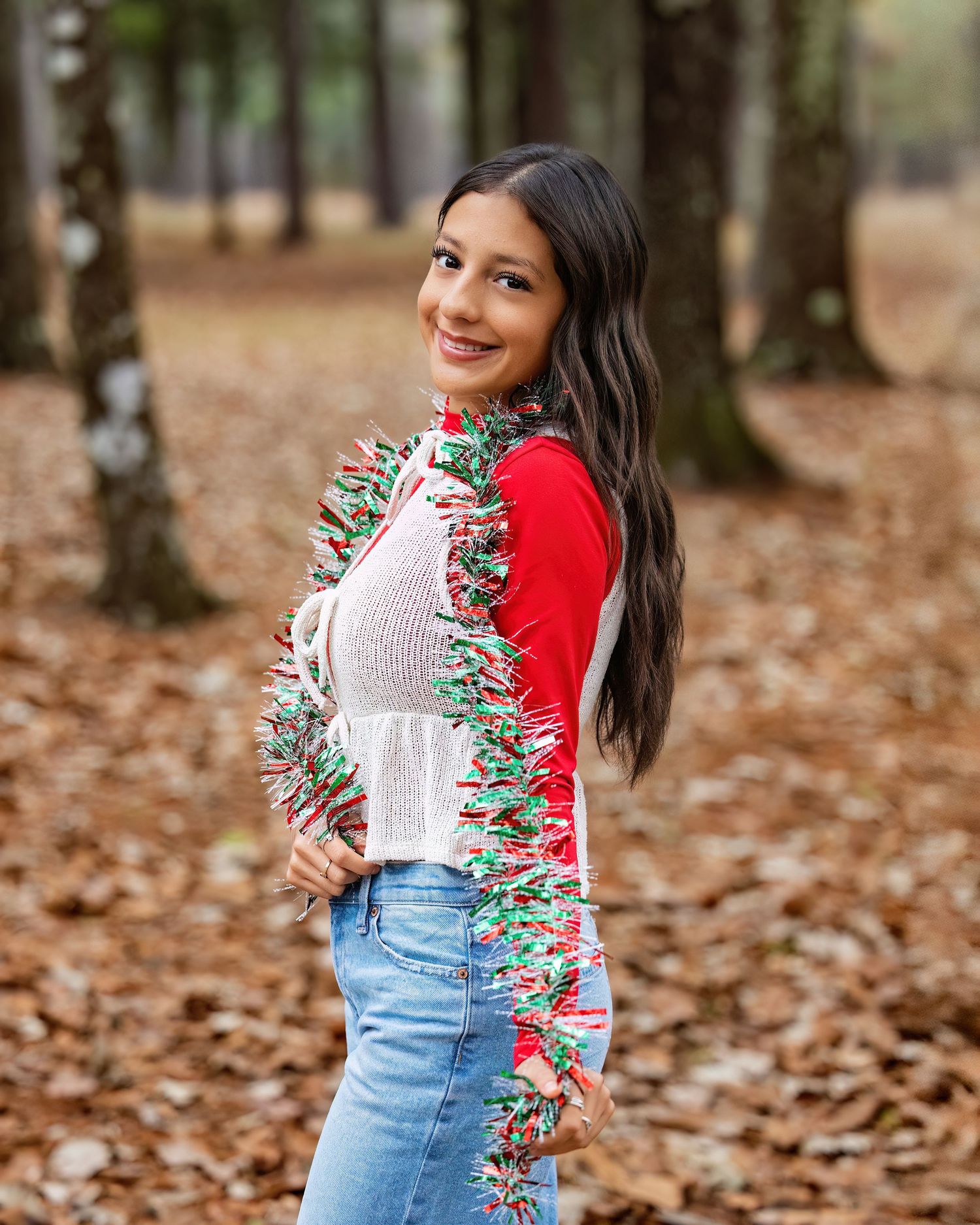 A woman in a colorful floral sweater and denim skirt poses playfully in a forest covered with fallen autumn leaves.