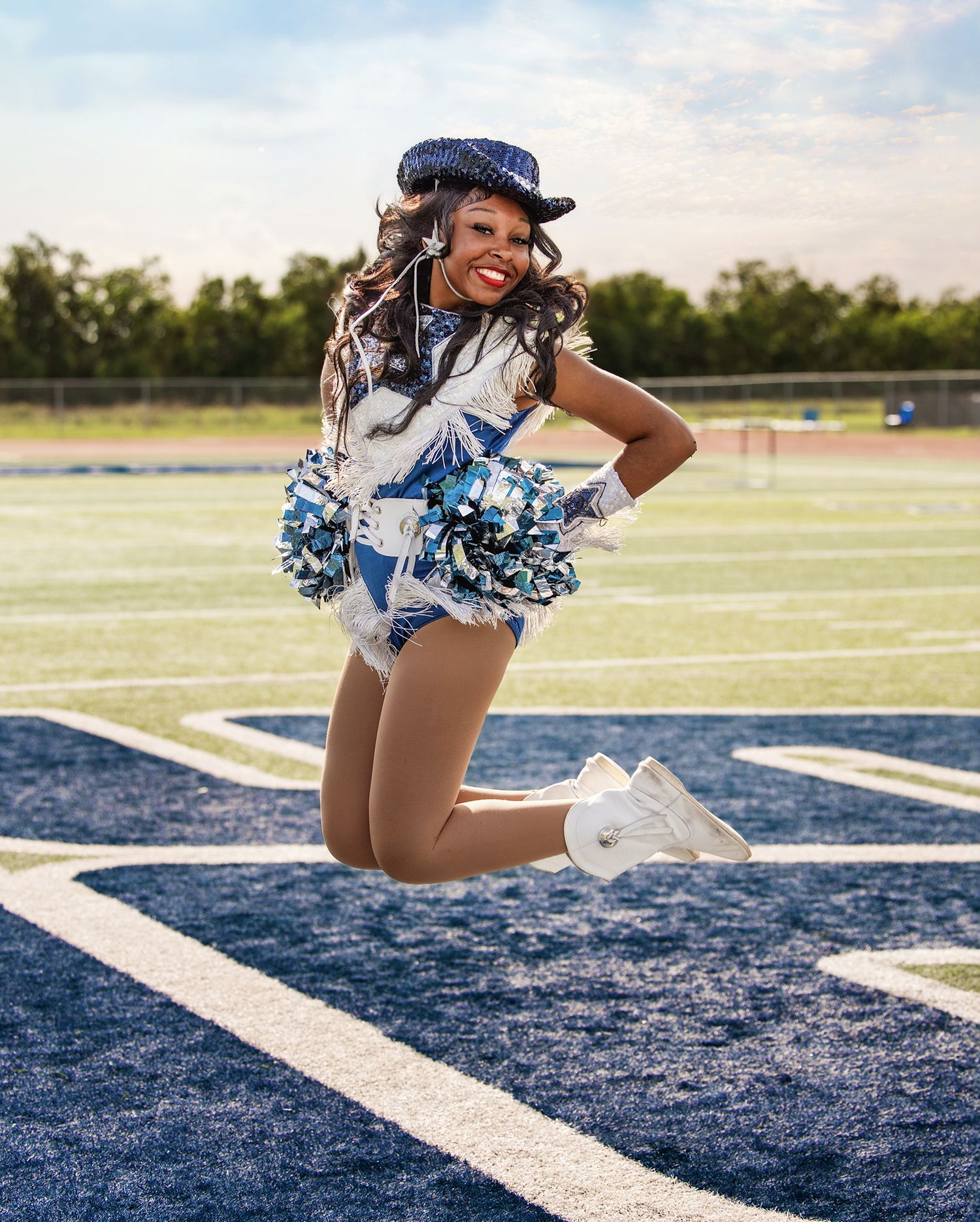 A cheerleader in blue and white uniform performs a dynamic pose on a football field with painted white lines.