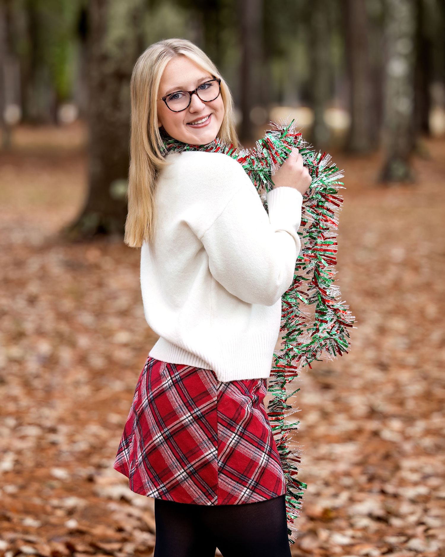 Person wearing glasses and white sweater holds holiday tinsel garland while standing among fallen autumn leaves outdoors.