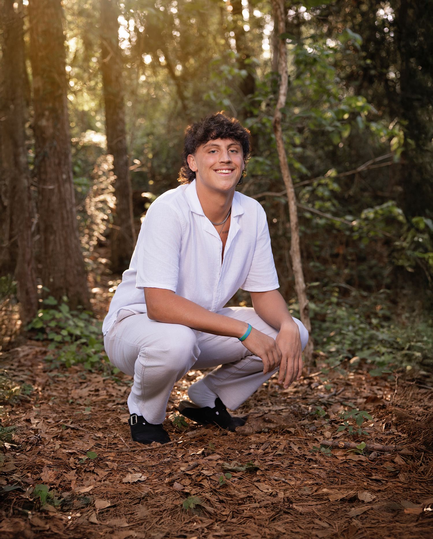 Young man in white shirt and gray pants crouching in a sunlit forest path, smiling at the camera.
