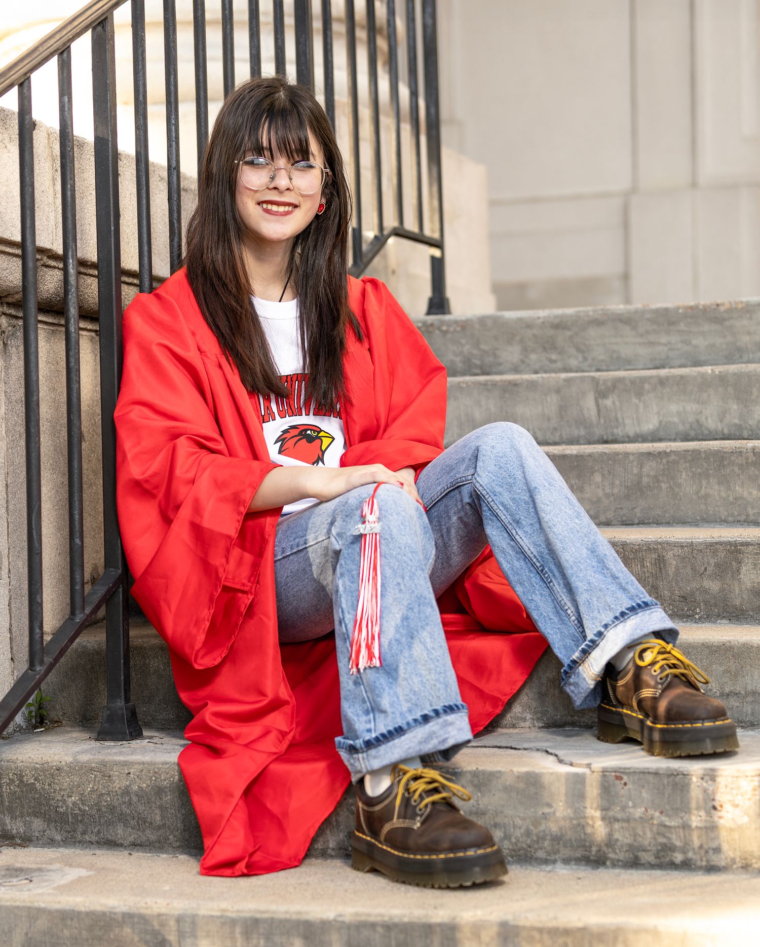 Person in bright red graduation gown sits on stone steps wearing casual jeans and brown boots.