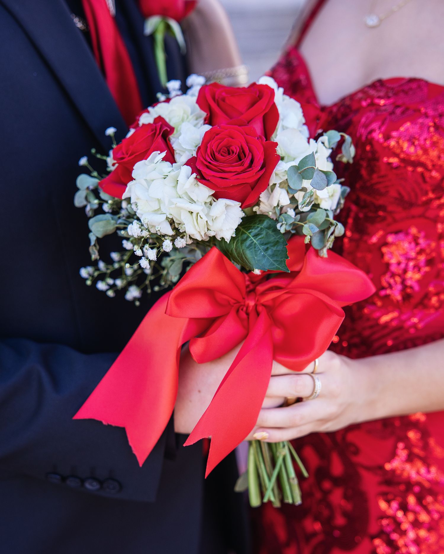 A red rose bouquet with white baby's breath flowers and a bright red satin bow accent against formal attire.