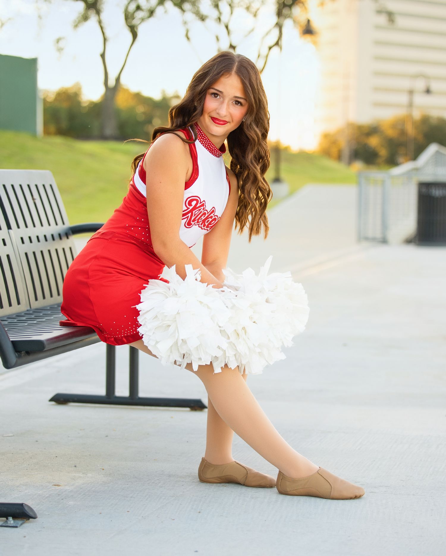 A cheerleader in red uniform performs dance poses on a concrete surface during golden hour lighting.