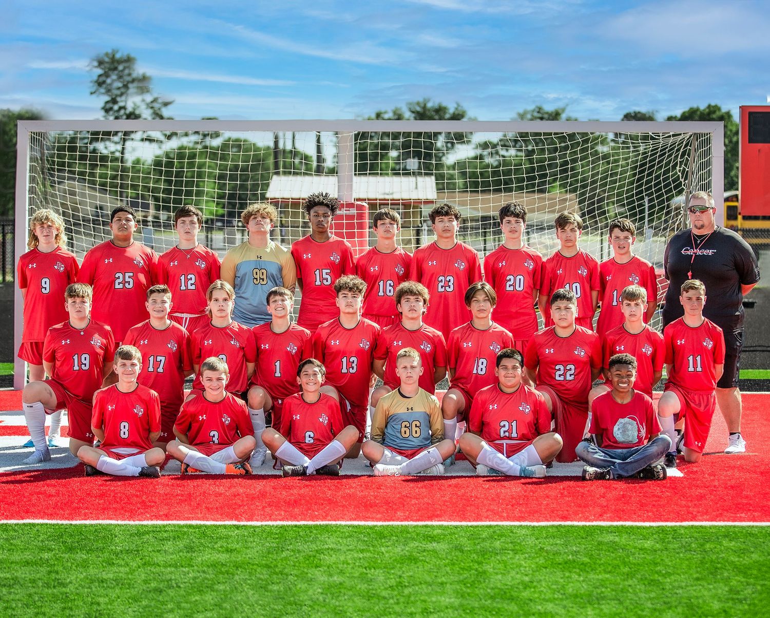 High school soccer team poses for group photo on red turf field with goal and blue sky in background.