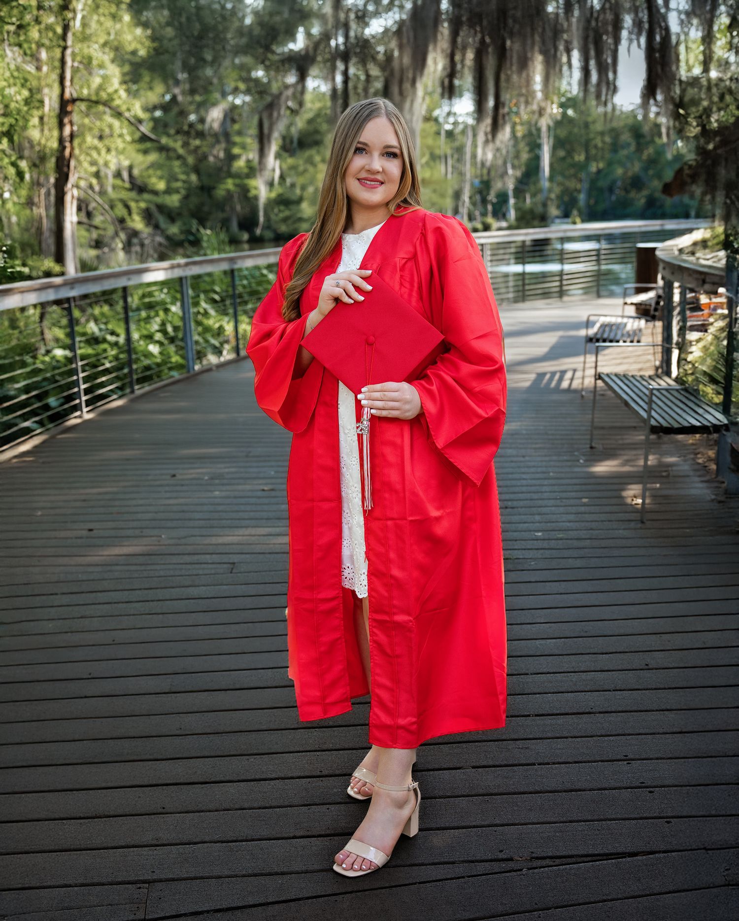 A person in a bright red graduation gown poses on a wooden deck walkway surrounded by lush greenery.