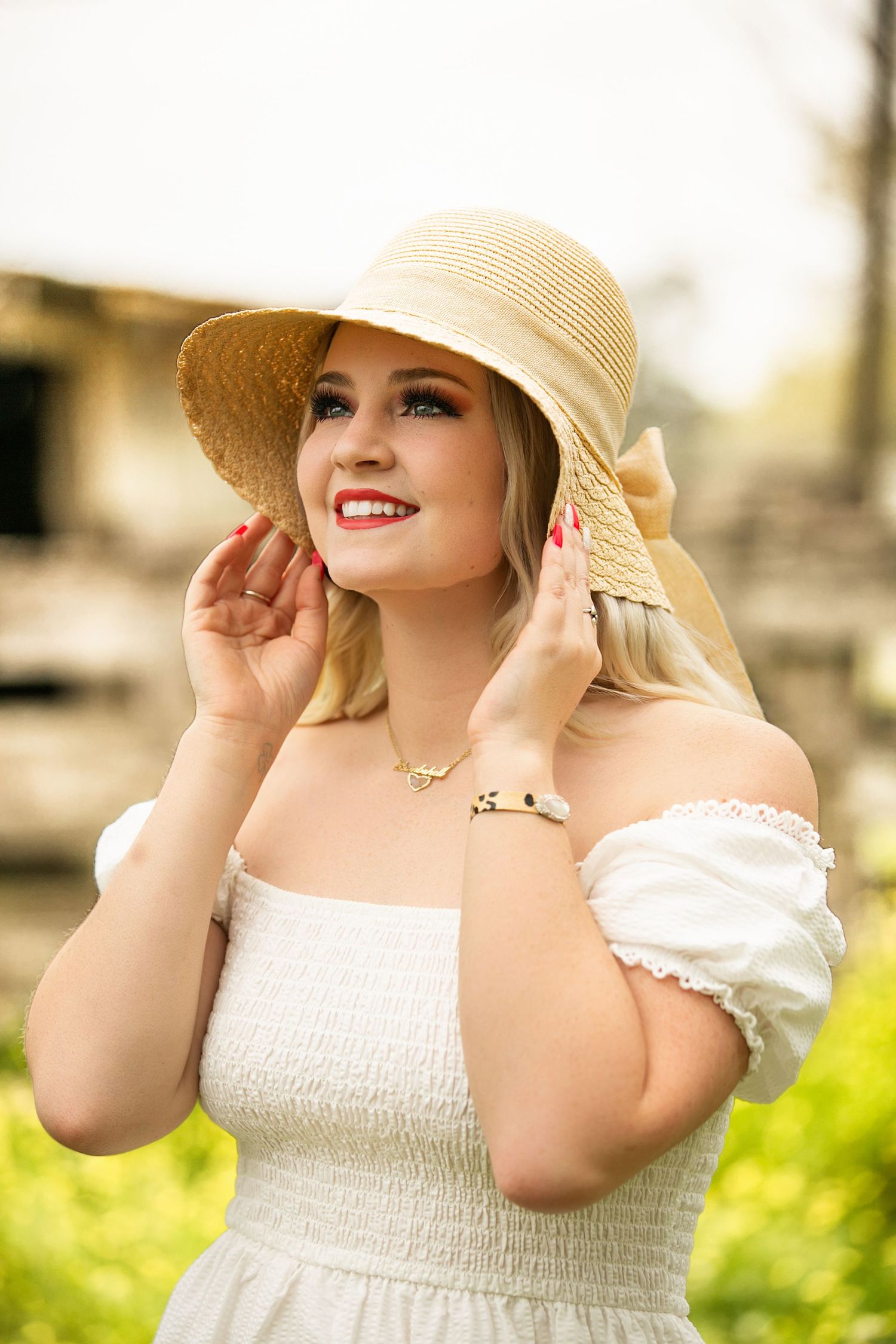 Smiling blonde woman in white off-shoulder dress and straw sun hat posing outdoors in a sunny garden setting.