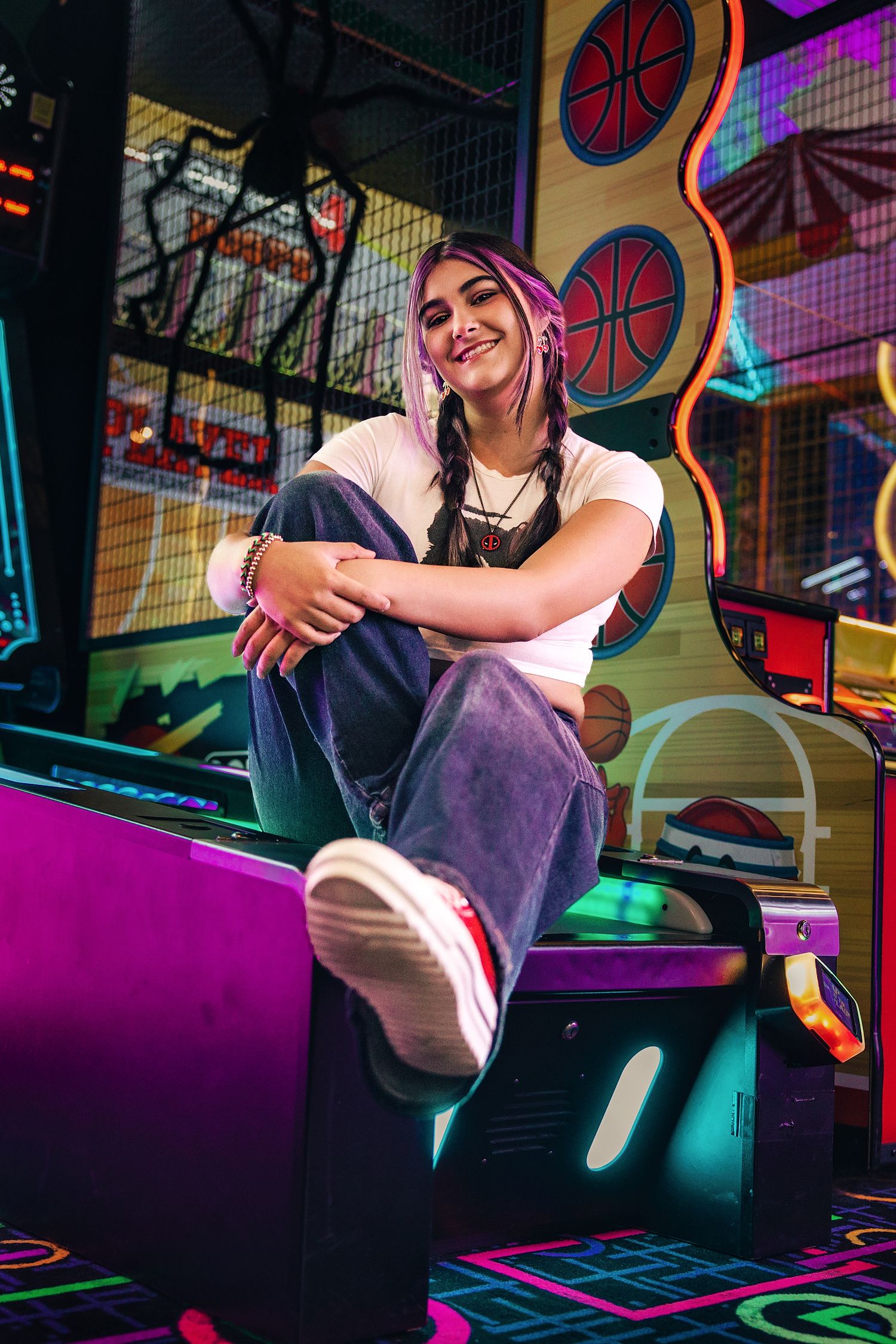 Young woman with braids sits smiling on arcade game in neon-lit entertainment center wearing white top and jeans.