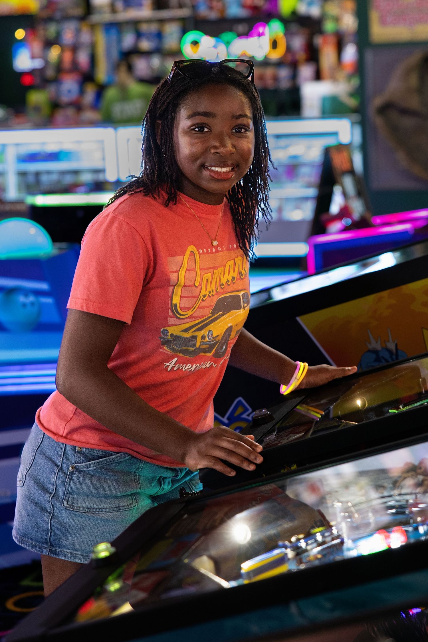 Young woman in red Camaro t-shirt smiling while playing arcade game in colorful game room.