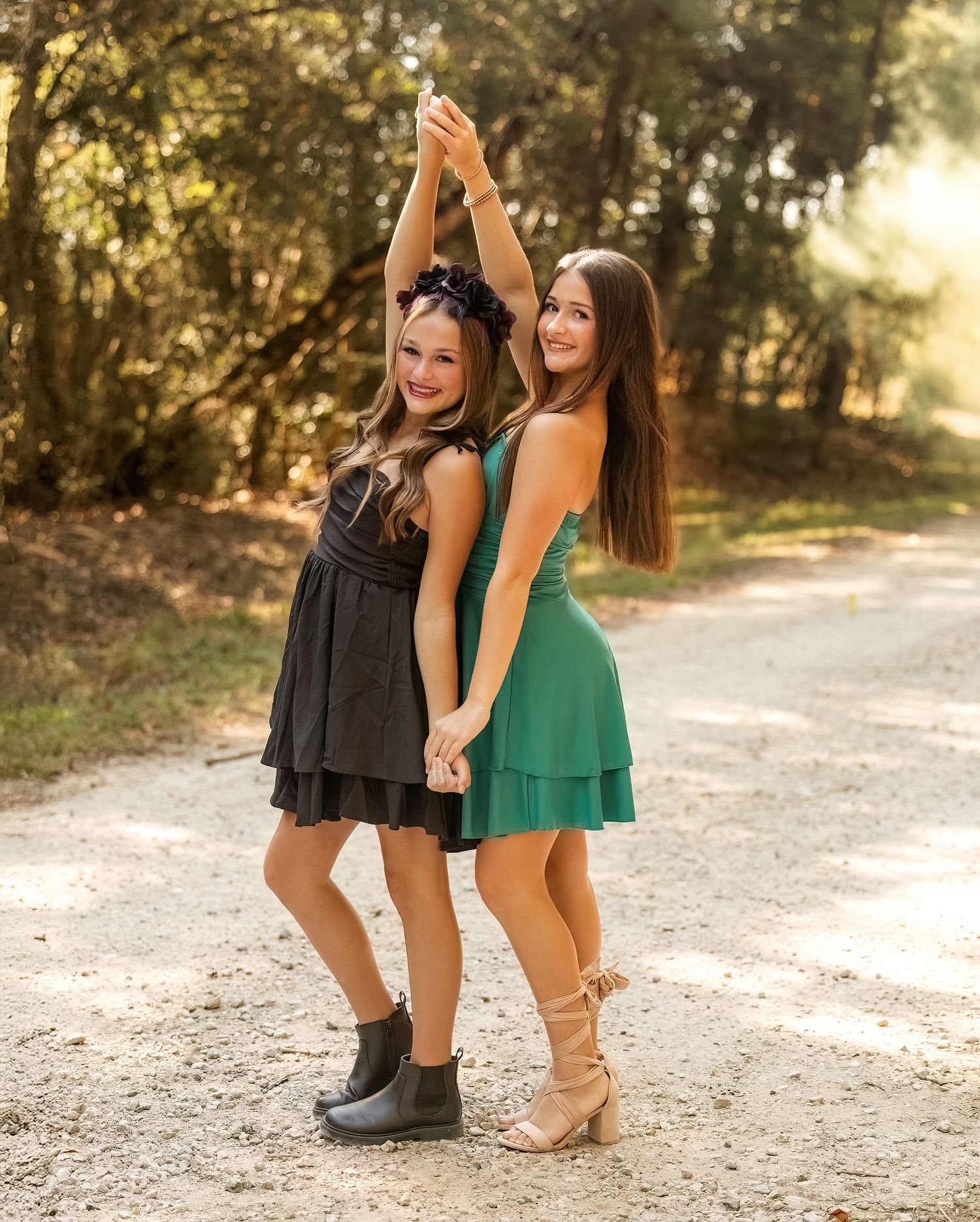 Two friends in contrasting black and green dresses pose back-to-back on a sunlit forest path. Best friend photography.