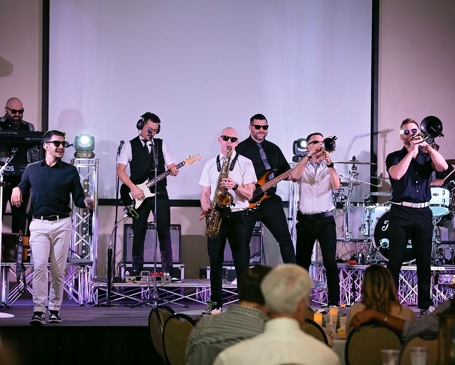 A concert live band performs on stage with multiple musicians playing instruments including saxophone, guitar and drums.