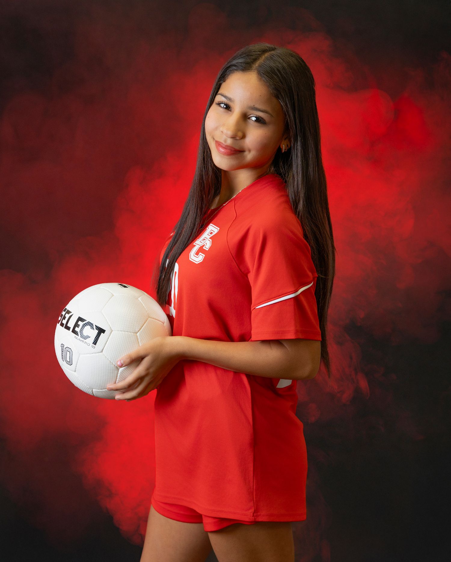 Youth volleyball player holds white ball while wearing red jersey against dark background with red lighting.