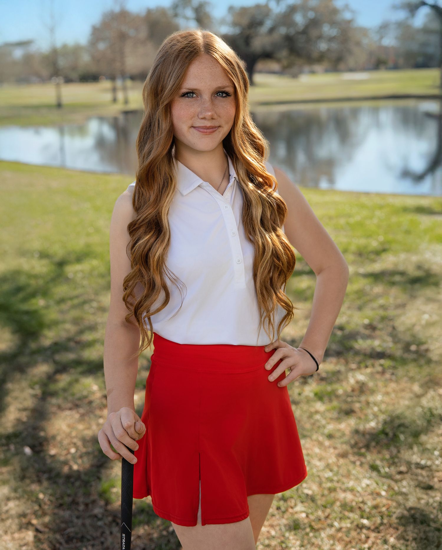 Young girl in white top and red shorts poses outdoors near a pond on a sunny day.