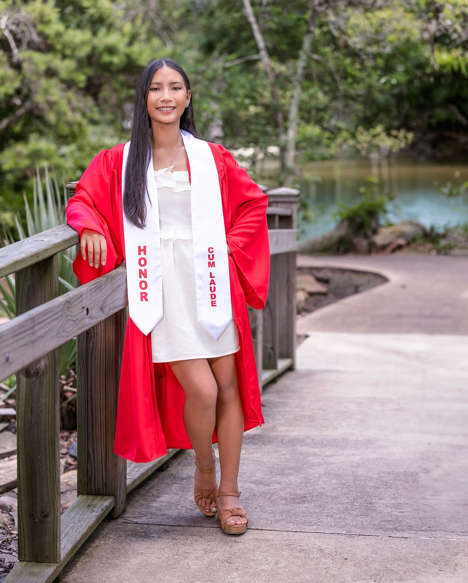 A person wearing a red blazer and white top poses outdoors on a wooden walkway near a lake.
