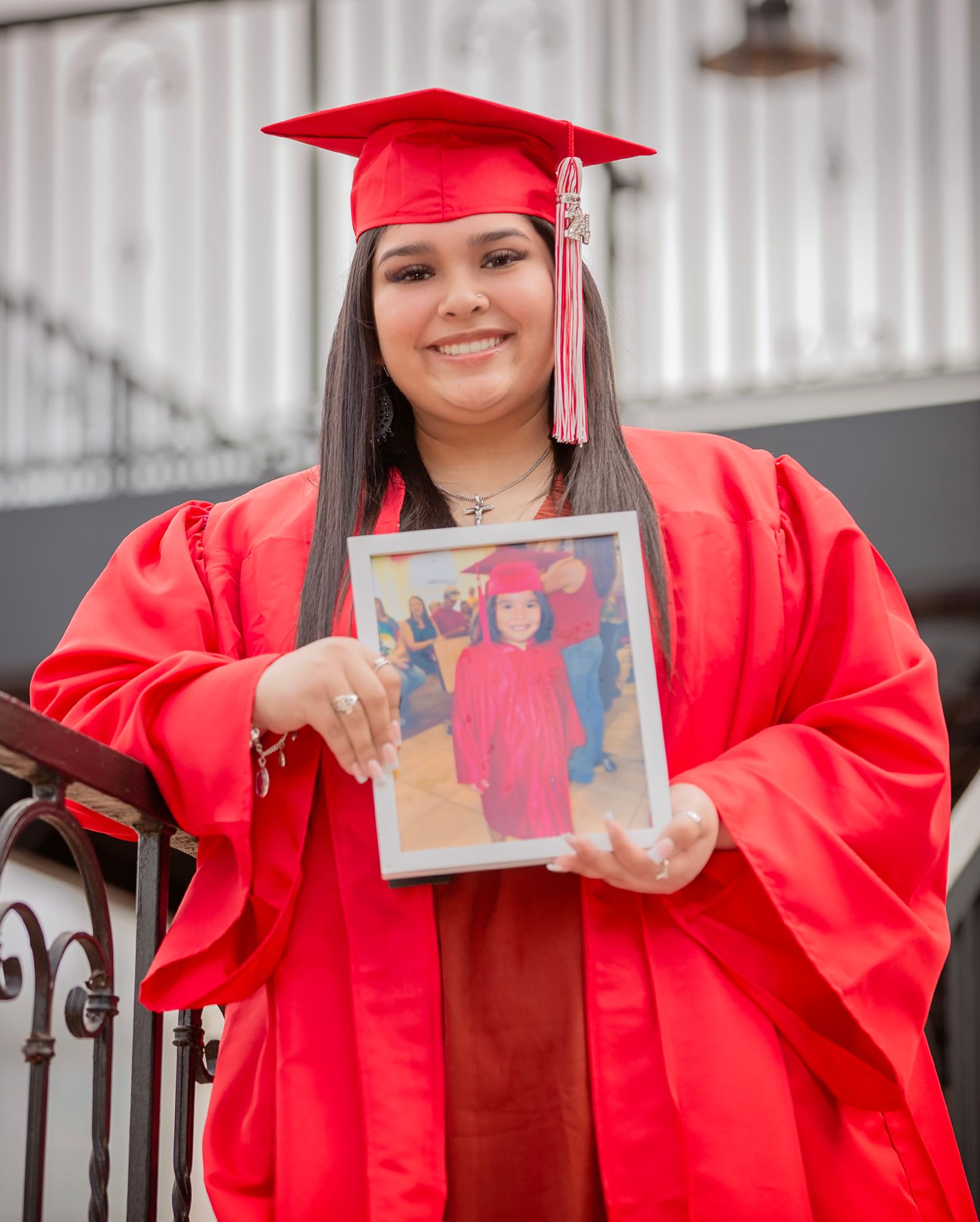Graduate in bright red cap and gown holds up a childhood graduation photo while standing on steps.