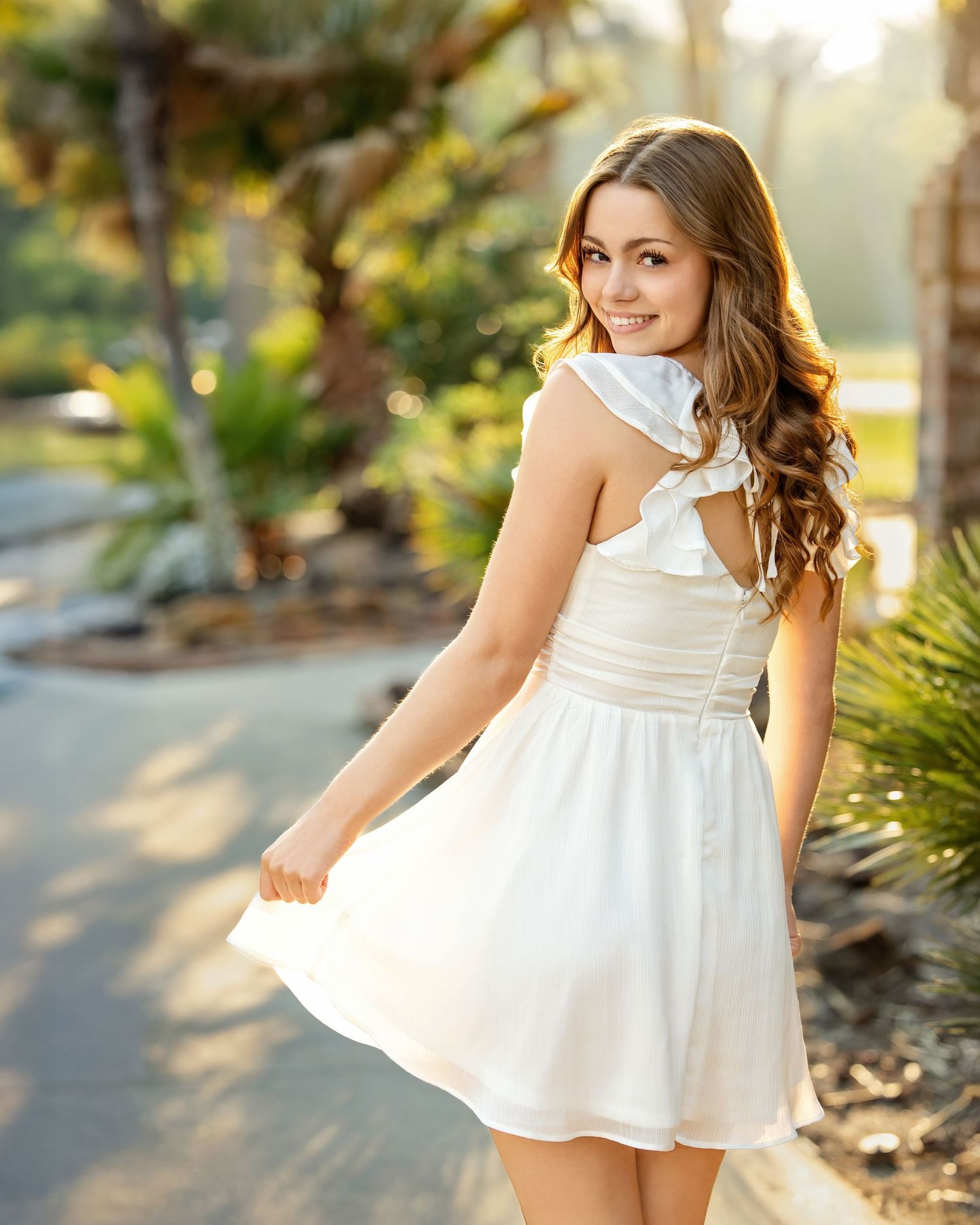 Young woman in white sundress smiling over shoulder on sunny garden path with palm trees.