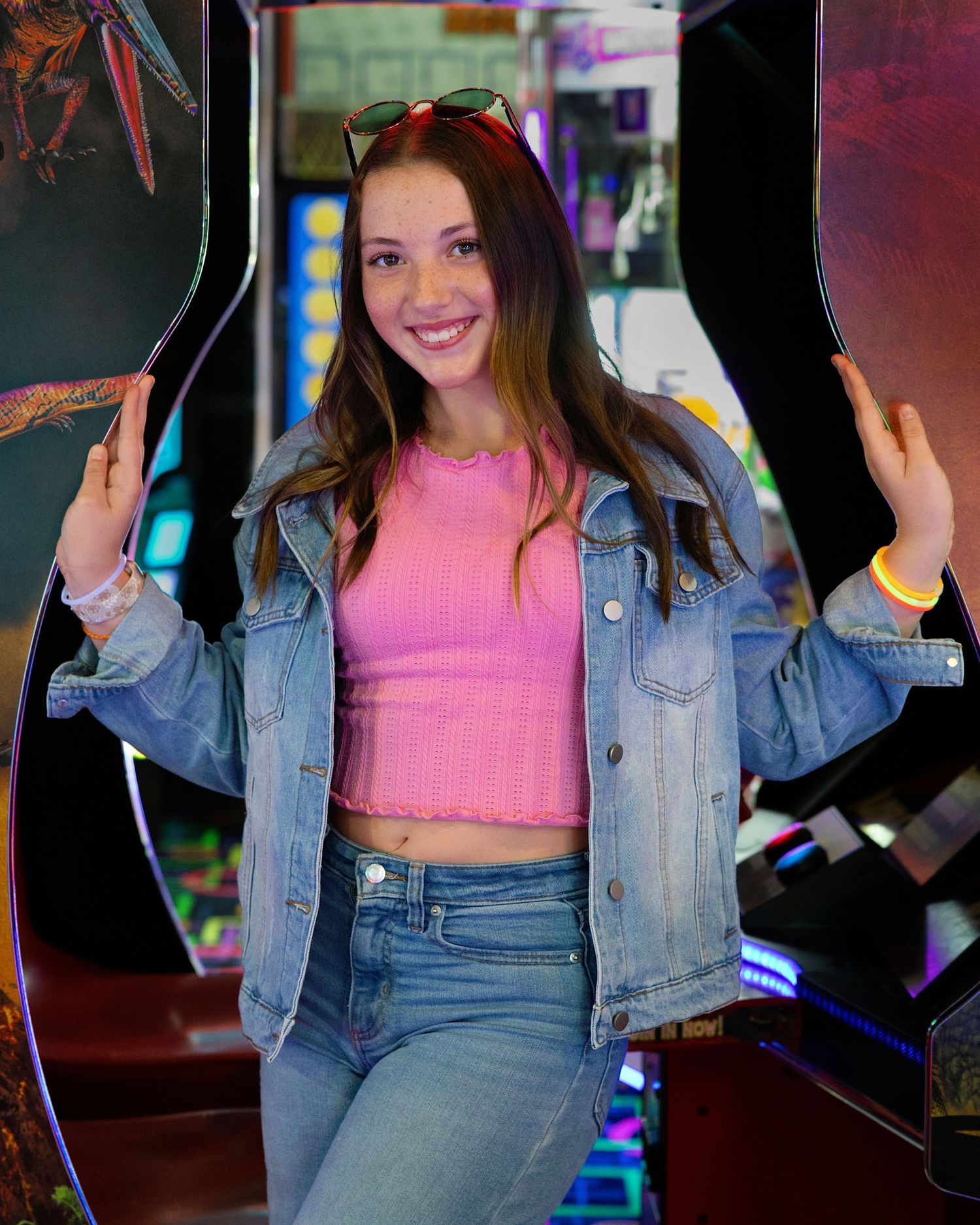 Smiling young woman in pink crop top and denim jacket posing between arcade machines in a game room.