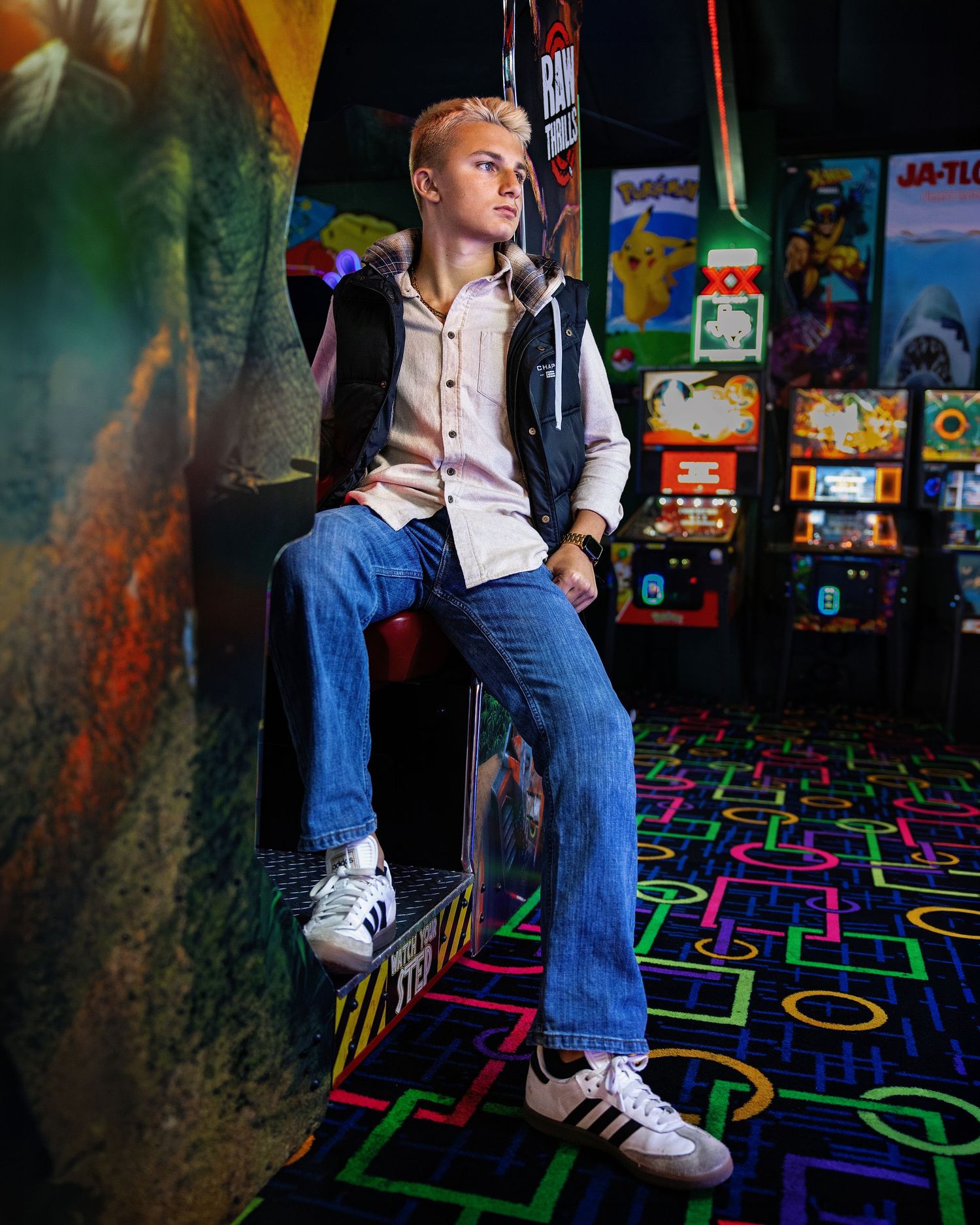 Young man in jeans and Adidas sneakers poses in a colorful retro arcade game room.