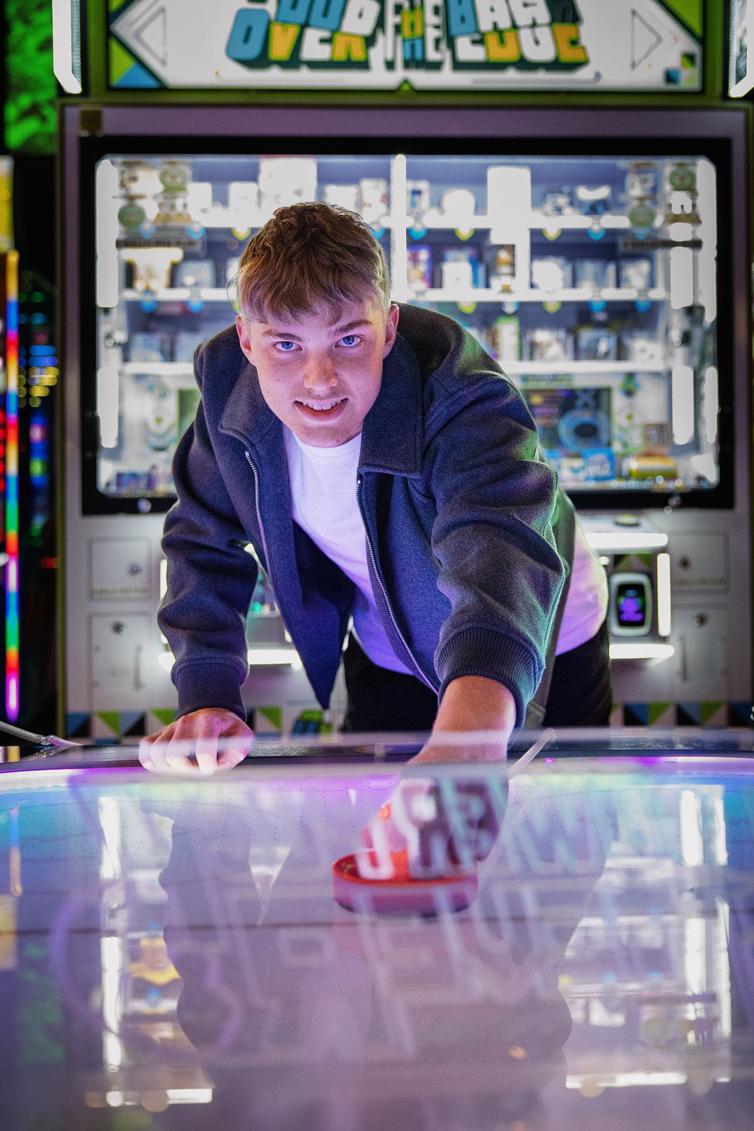 Young man playing air hockey at an arcade, leaning over the table with intensity, colorful prize machines in background.