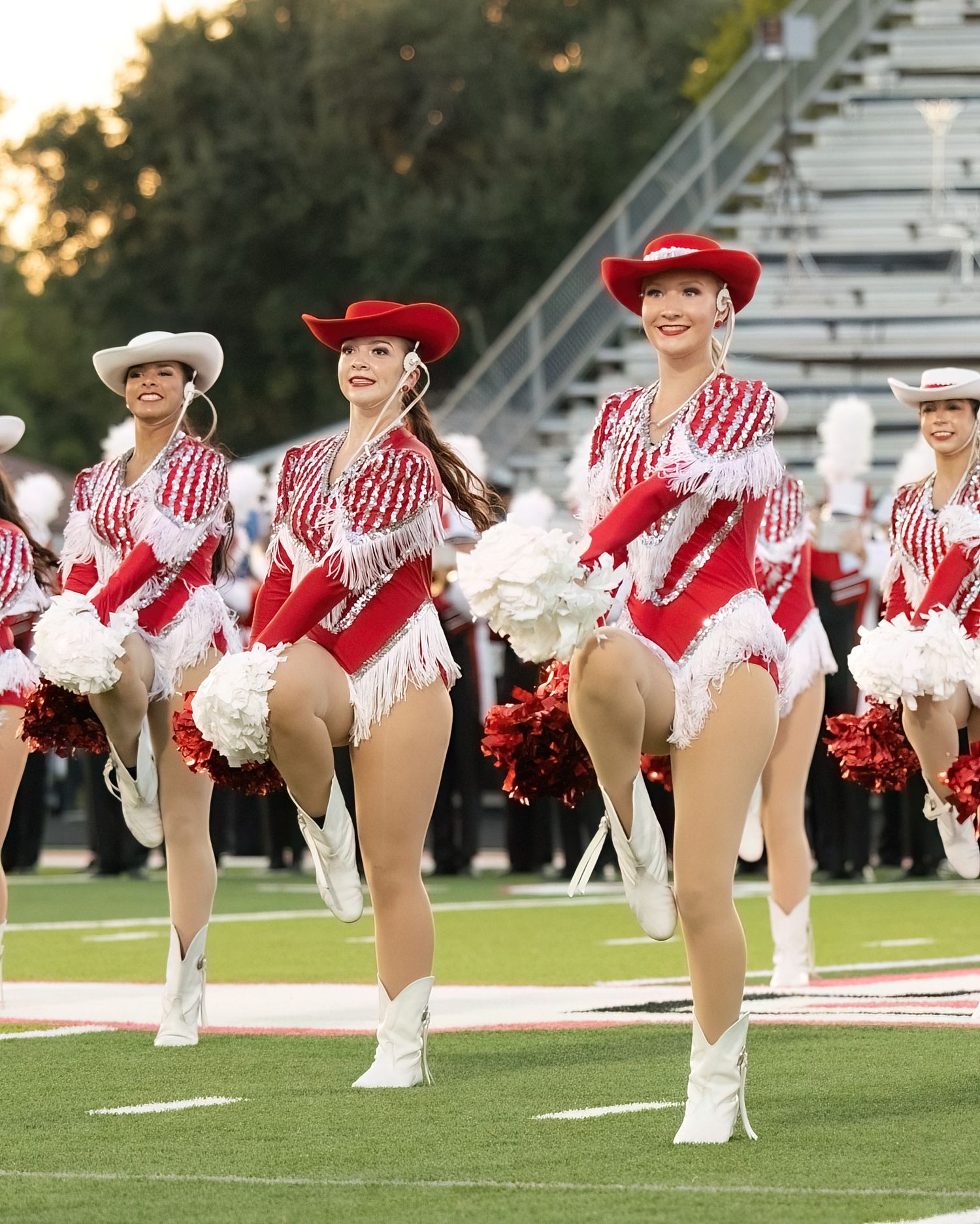 Dance team members in red and white uniforms perform synchronized high kicks on a football field at sunset.