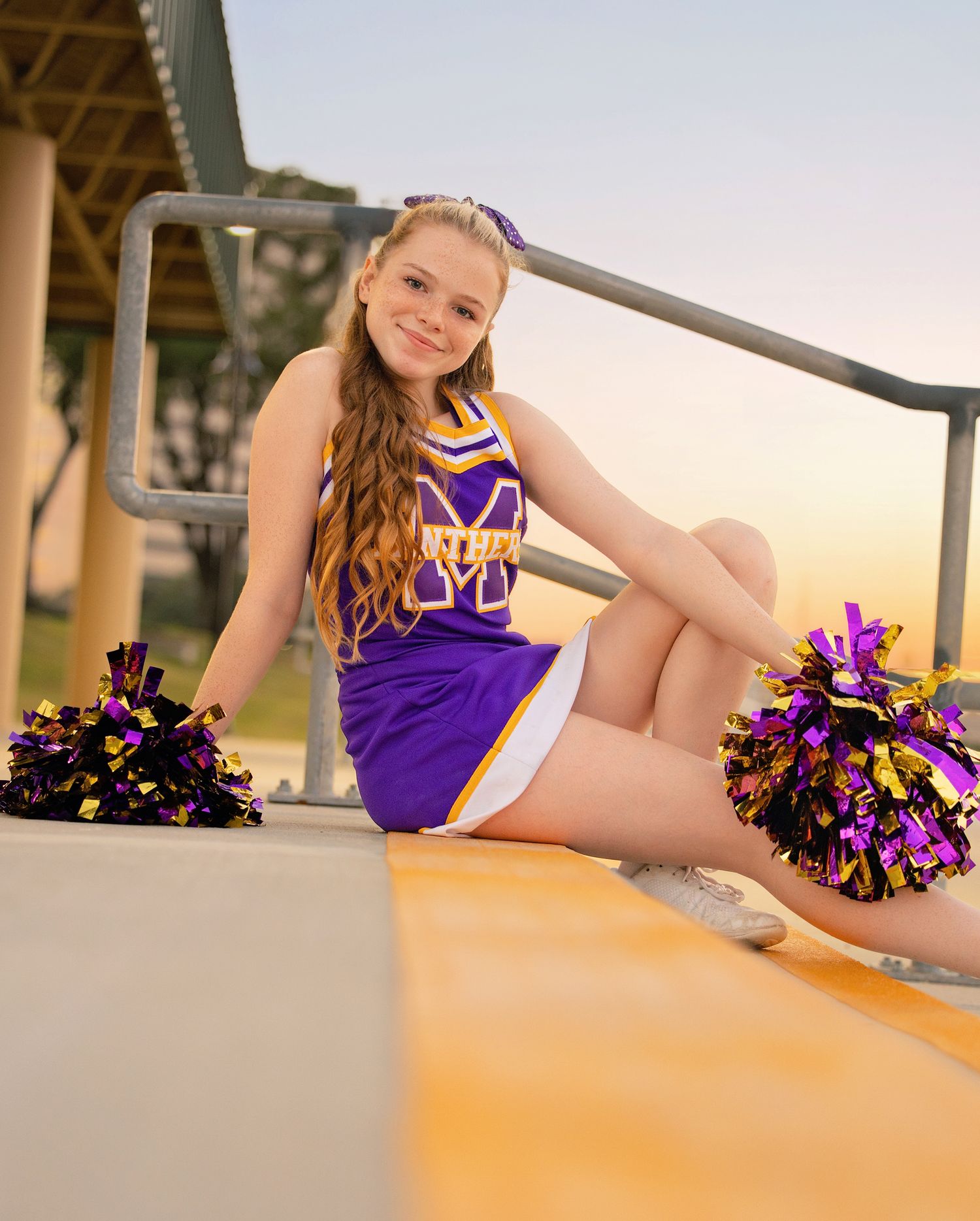 Cheerleader in purple uniform posing with pom poms on yellow surface during outdoor photoshoot.