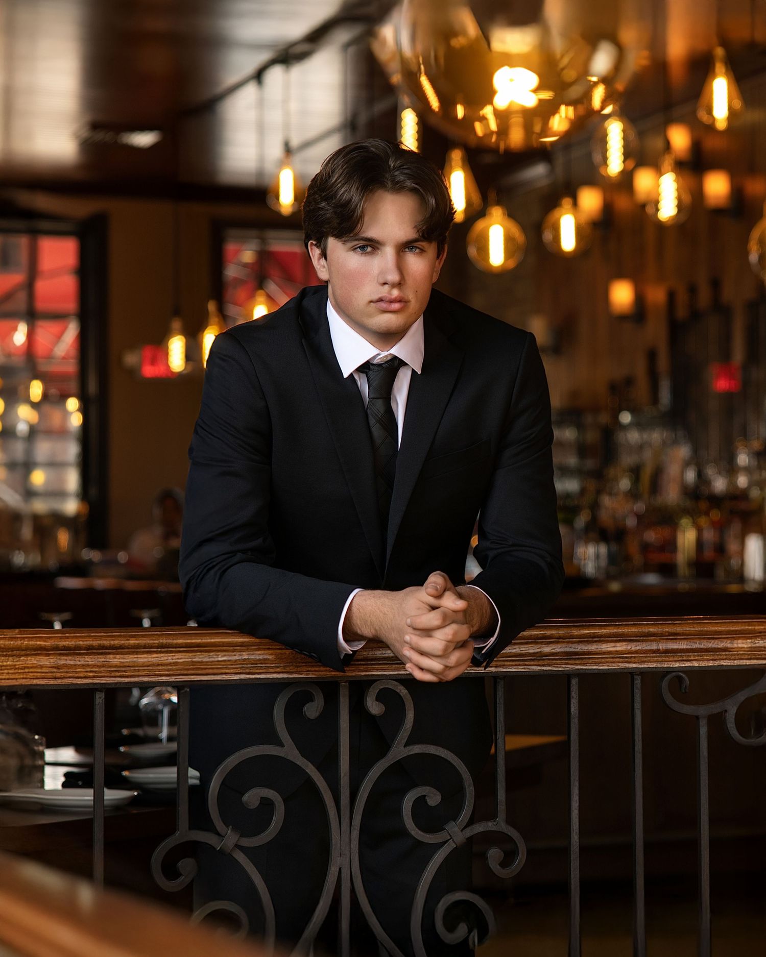 Young man in black suit and tie leaning on a bar counter in an upscale restaurant with warm Edison bulb lighting.