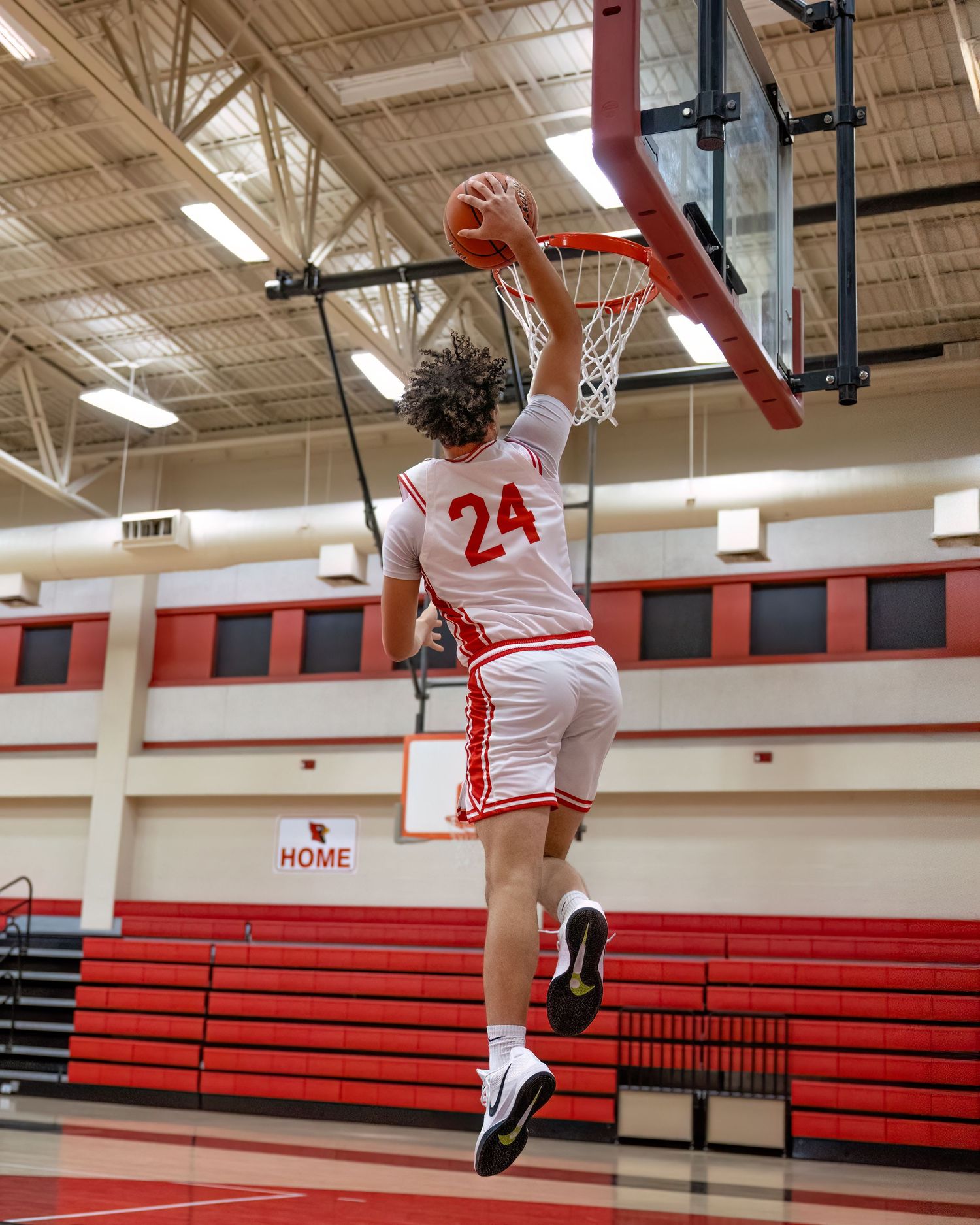 Basketball player in white jersey number 24 performs an athletic layup in a high school gymnasium with red bleachers.