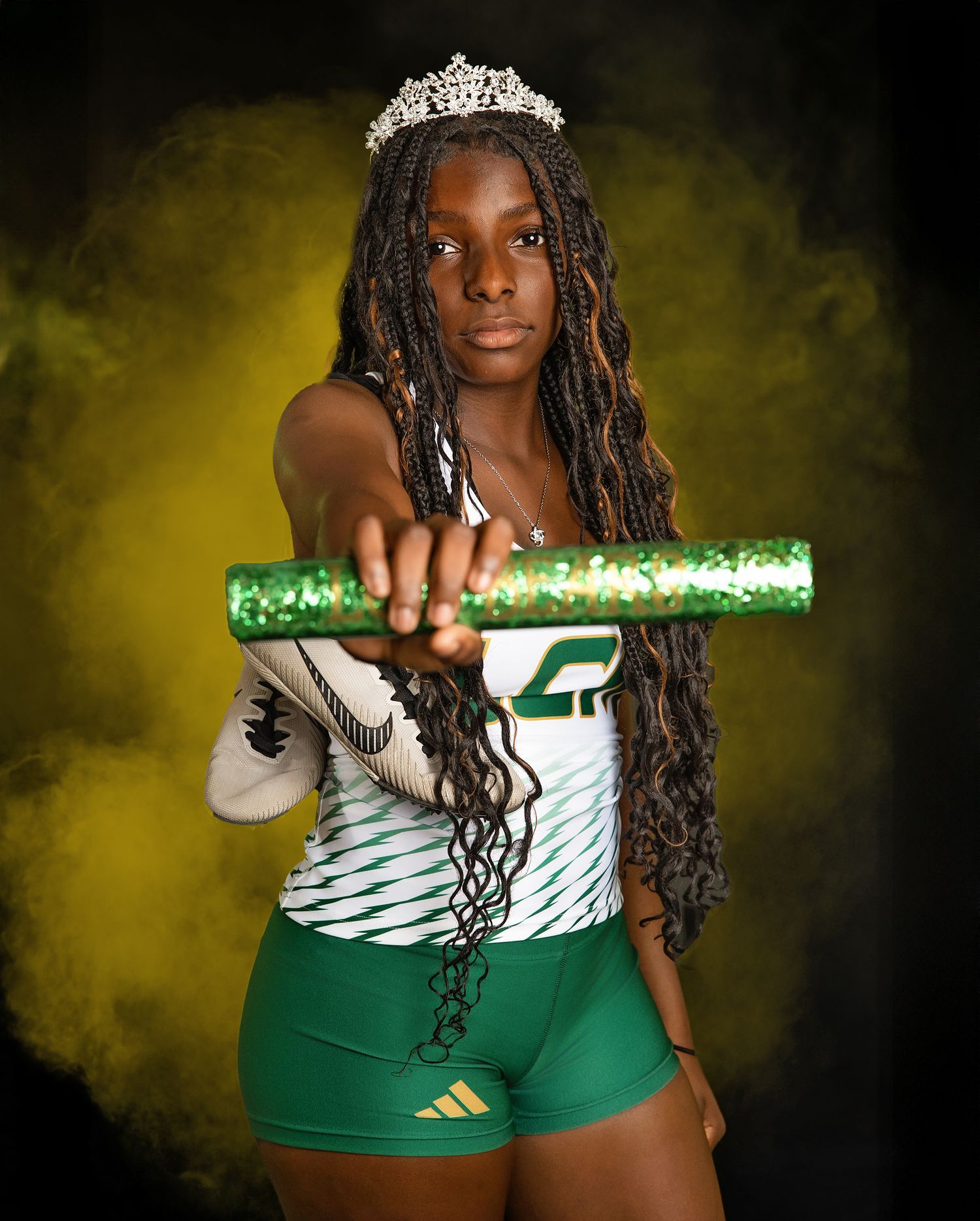 Athlete in green and white uniform holds a sparkly baton while wearing a tiara against a dramatic yellow background.
