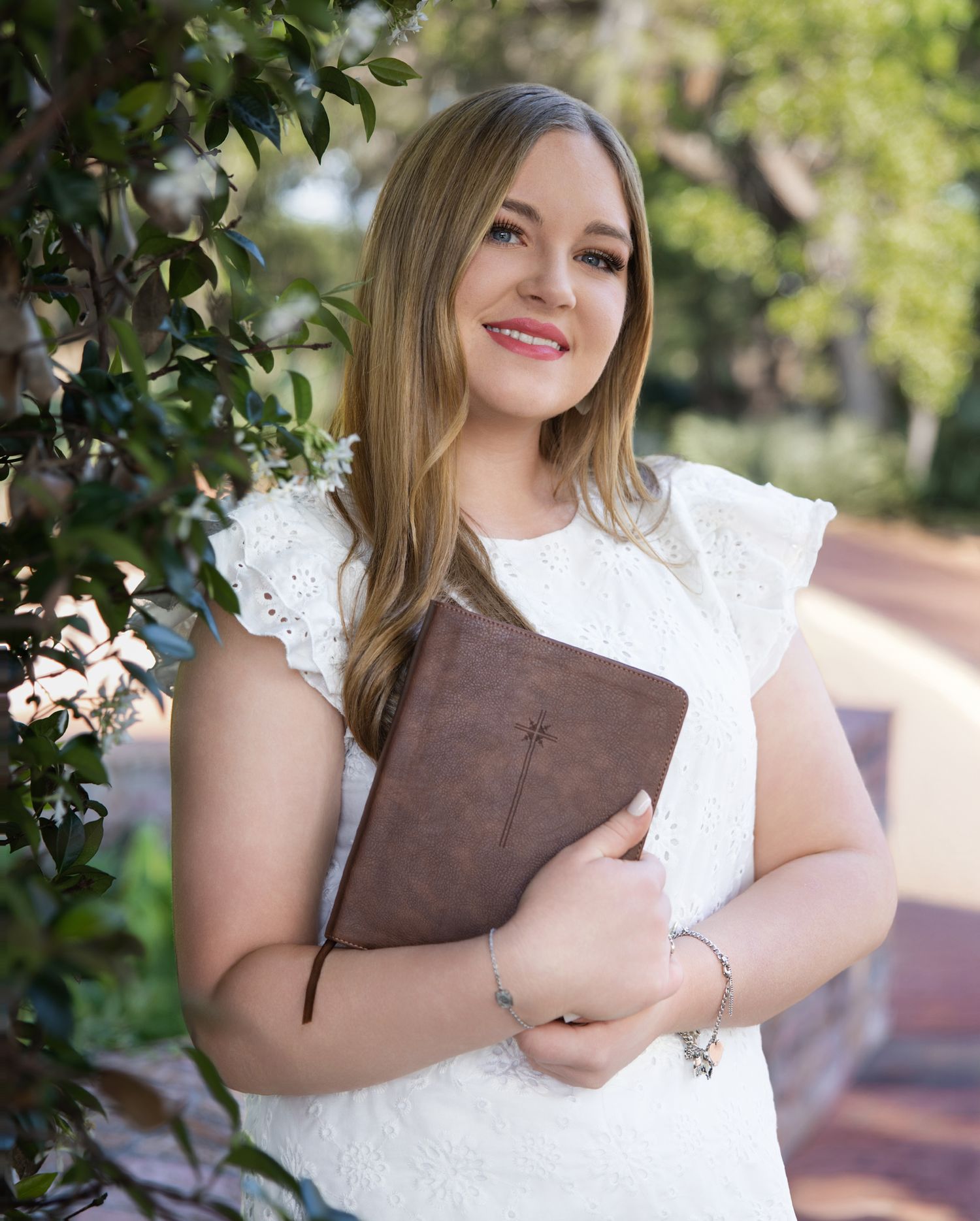 A person in a white lace dress holds a brown leather book while standing outdoors near green foliage.