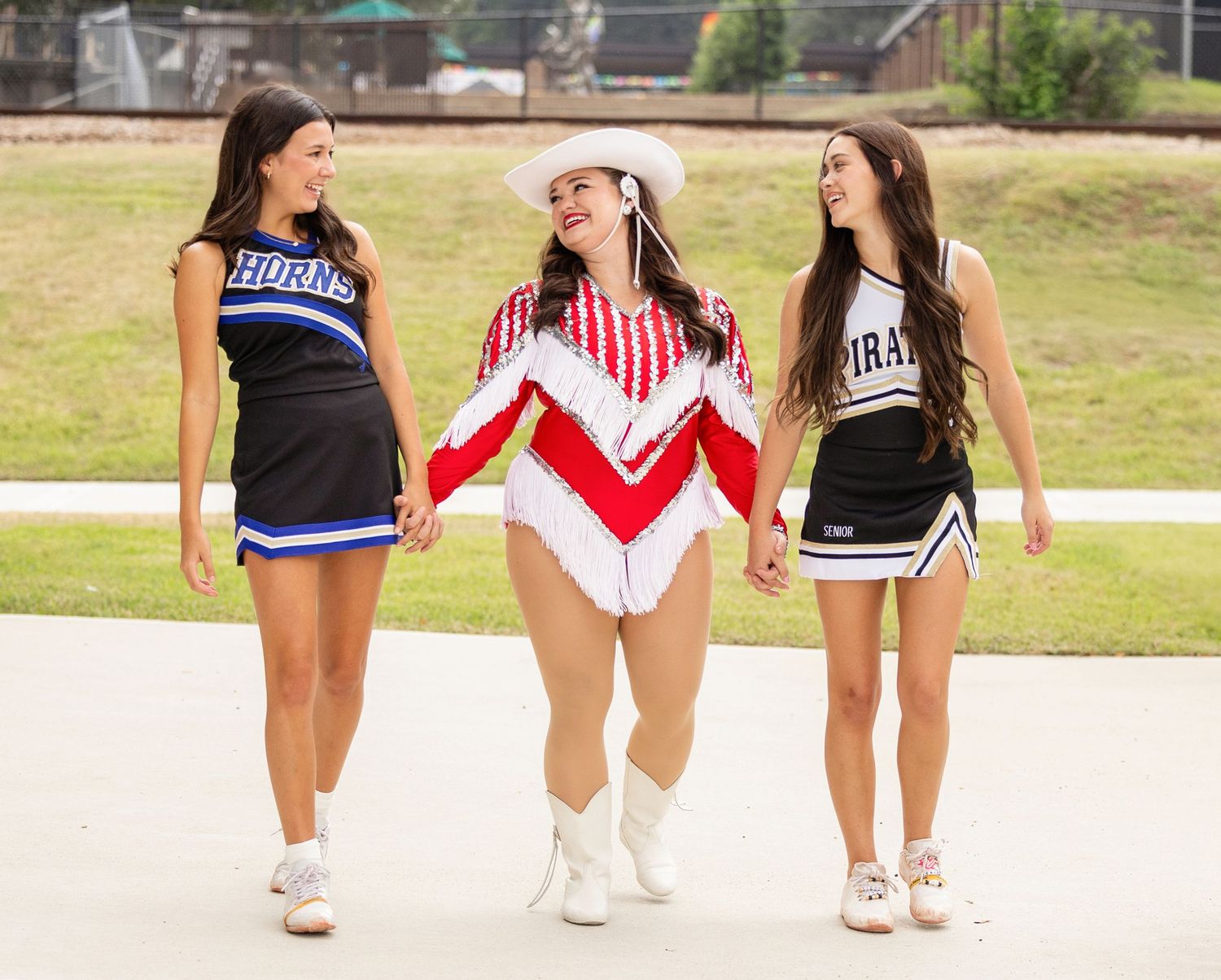 Three cheerleaders in black and white uniforms sit together on a white ledge outdoors.