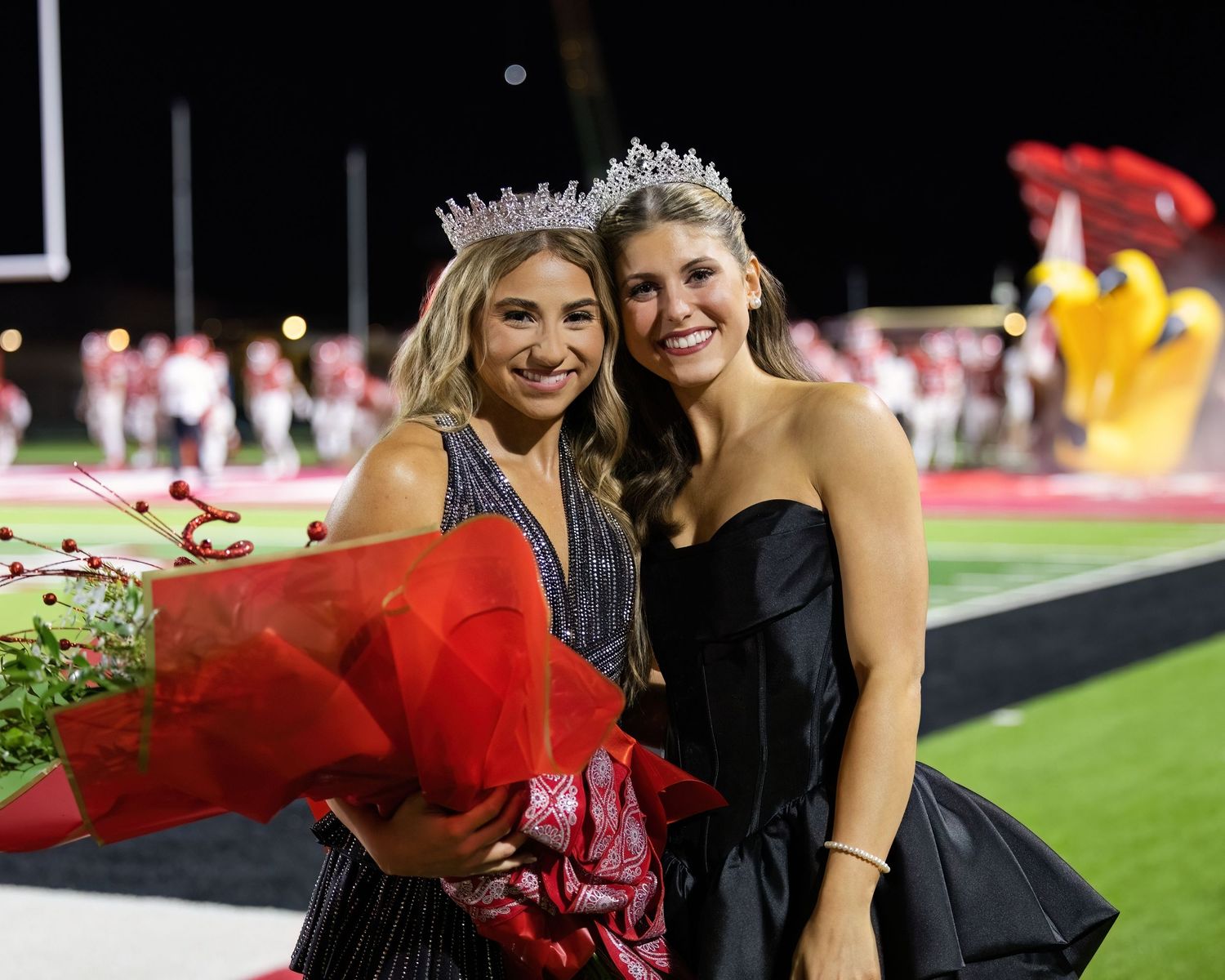People pose together on football field at night holding red bouquets.