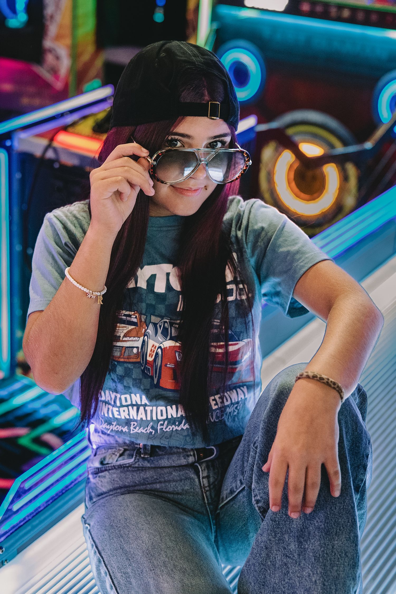 Young woman in Daytona International Speedway tee, cap, and aviator sunglasses posing in front of neon arcade lights.