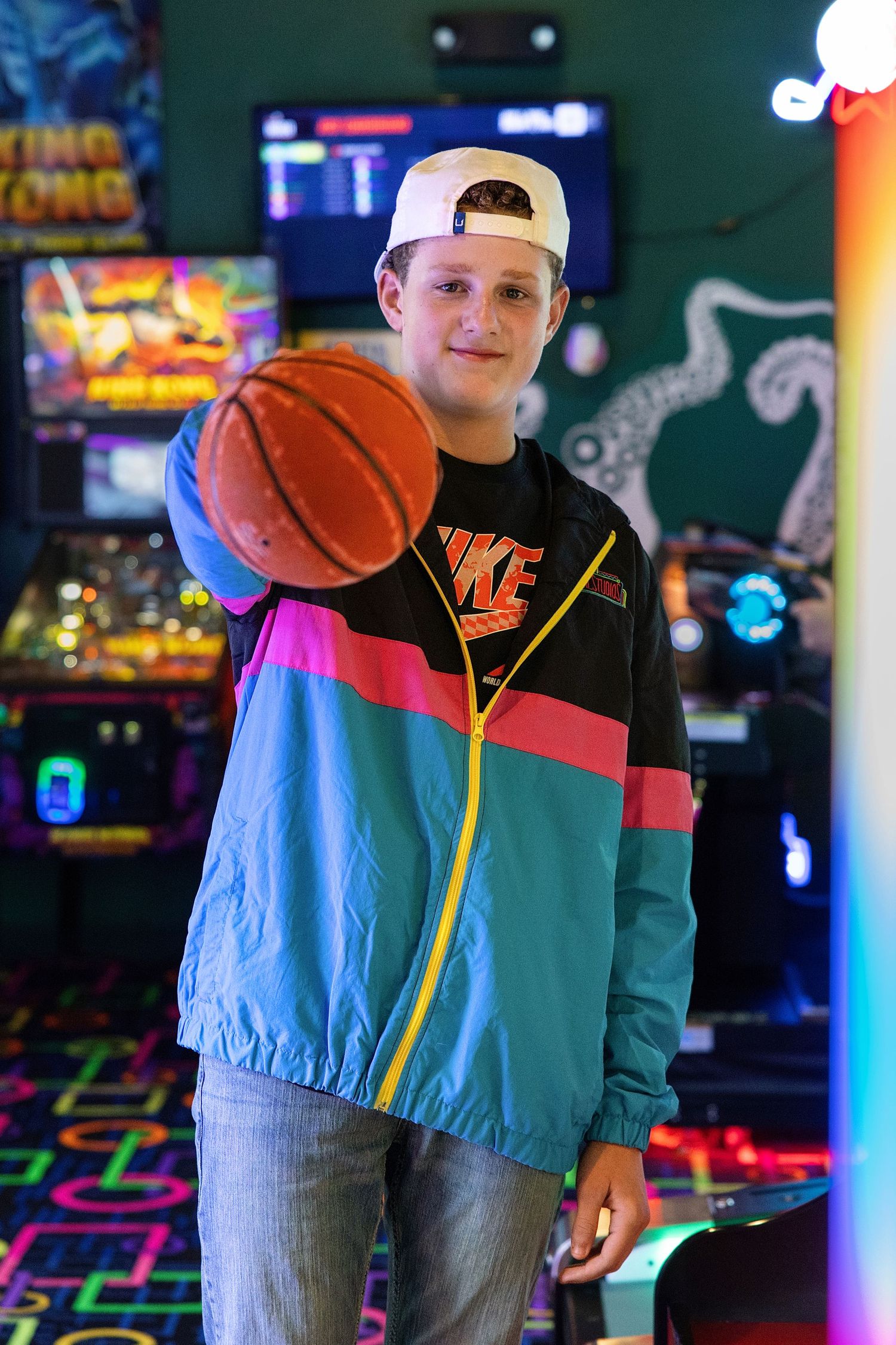 Teen boy in retro windbreaker and backwards cap holds basketball in colorful arcade game room.