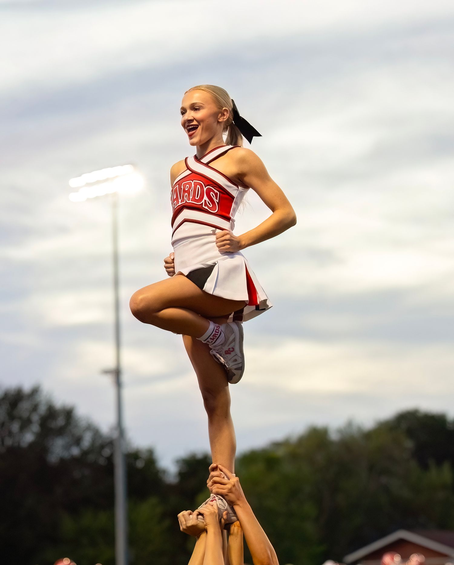 A cheerleader performs a high liberty stunt against a cloudy sky during an outdoor performance.