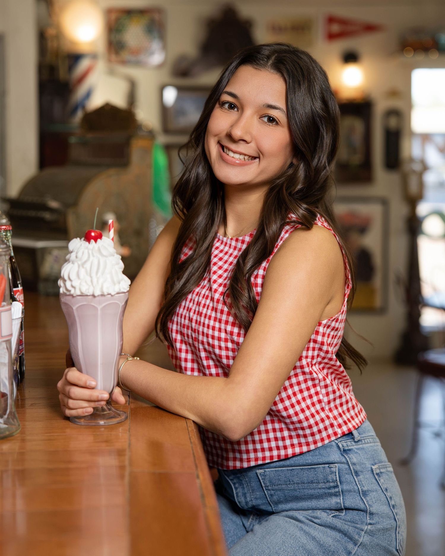 Person in red gingham top and jeans enjoys milkshake at retro diner counter with vintage decor in background.
