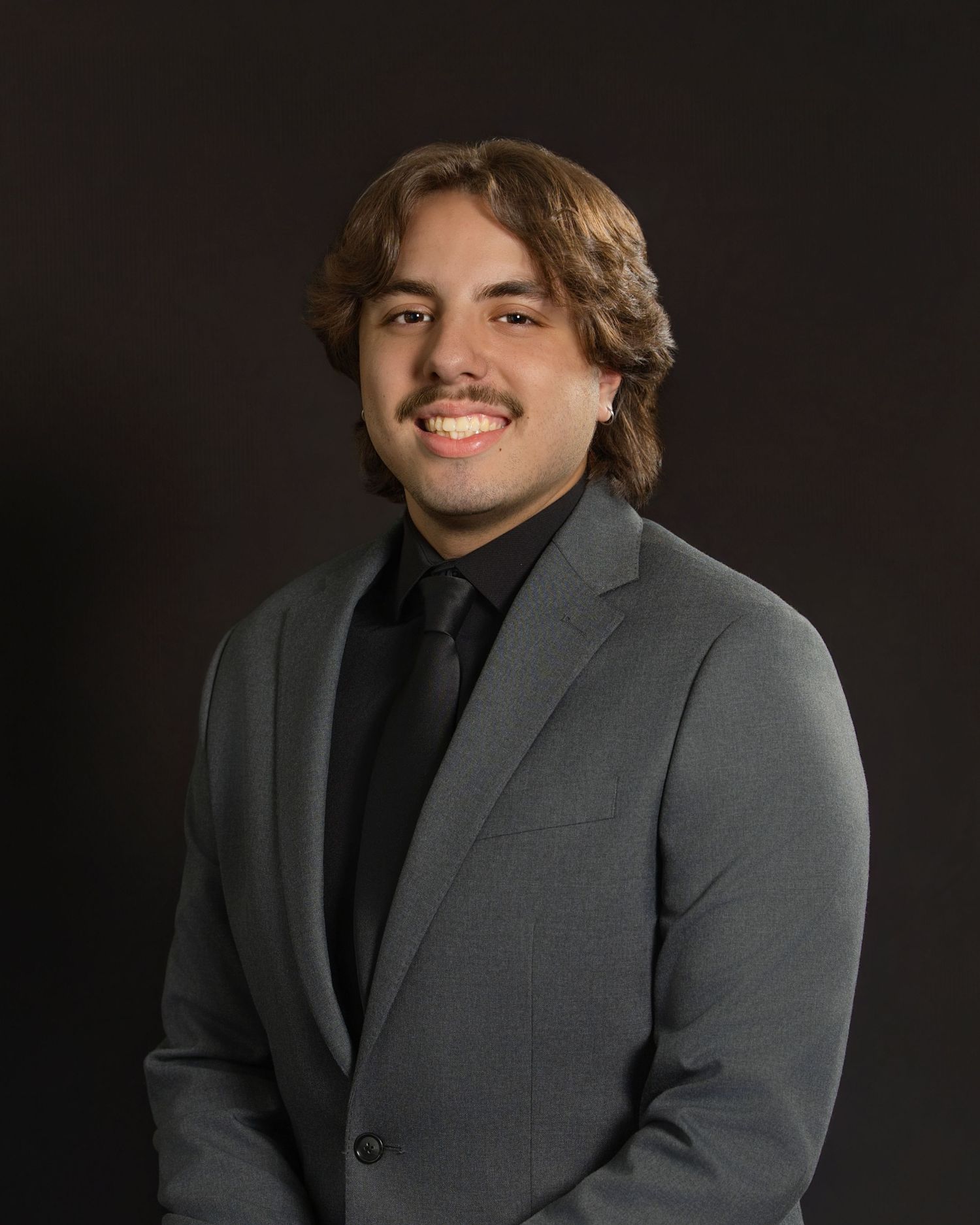 Professional headshot of someone in a gray suit jacket against a dark background smiling at the camera.