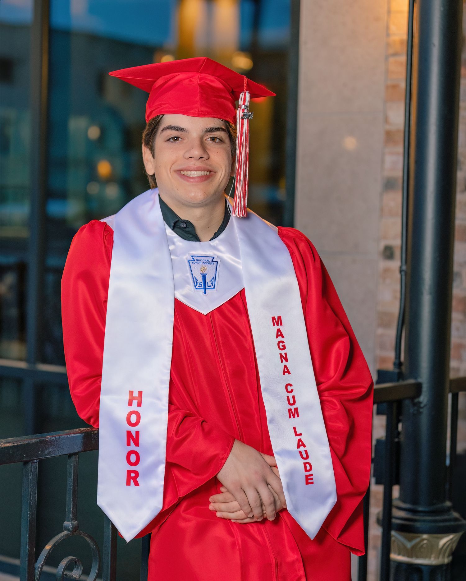 A group of graduates wearing red caps and gowns with white sashes stand together outside a building.