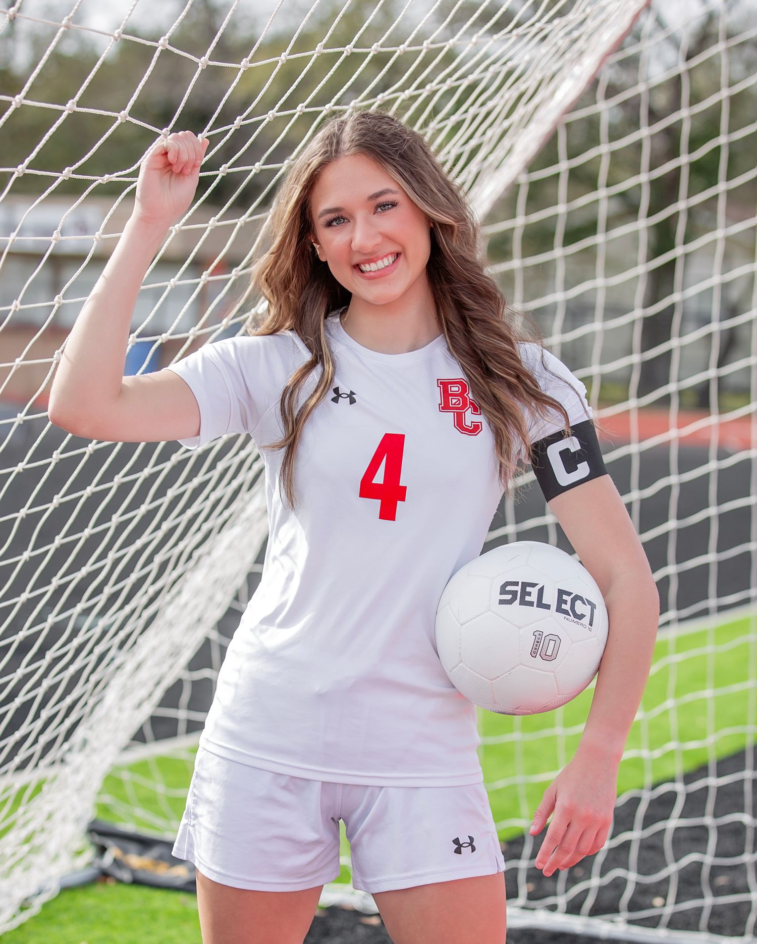 Soccer player in white uniform with number 4 poses with ball by goal net on sports field.