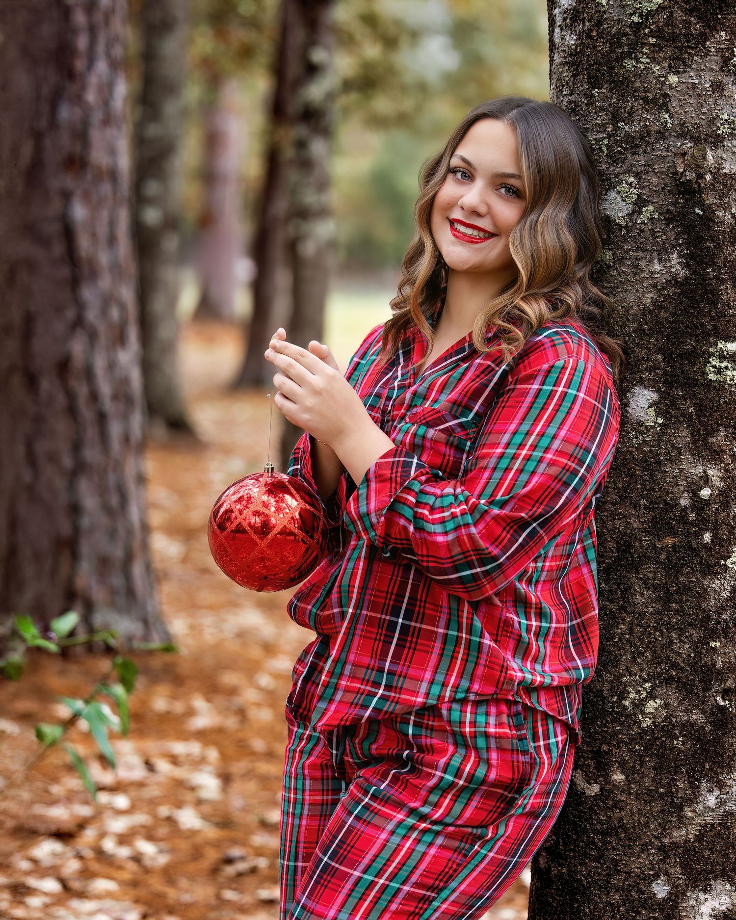 Someone in a red plaid shirt poses playfully against a tree trunk in an autumn forest setting.