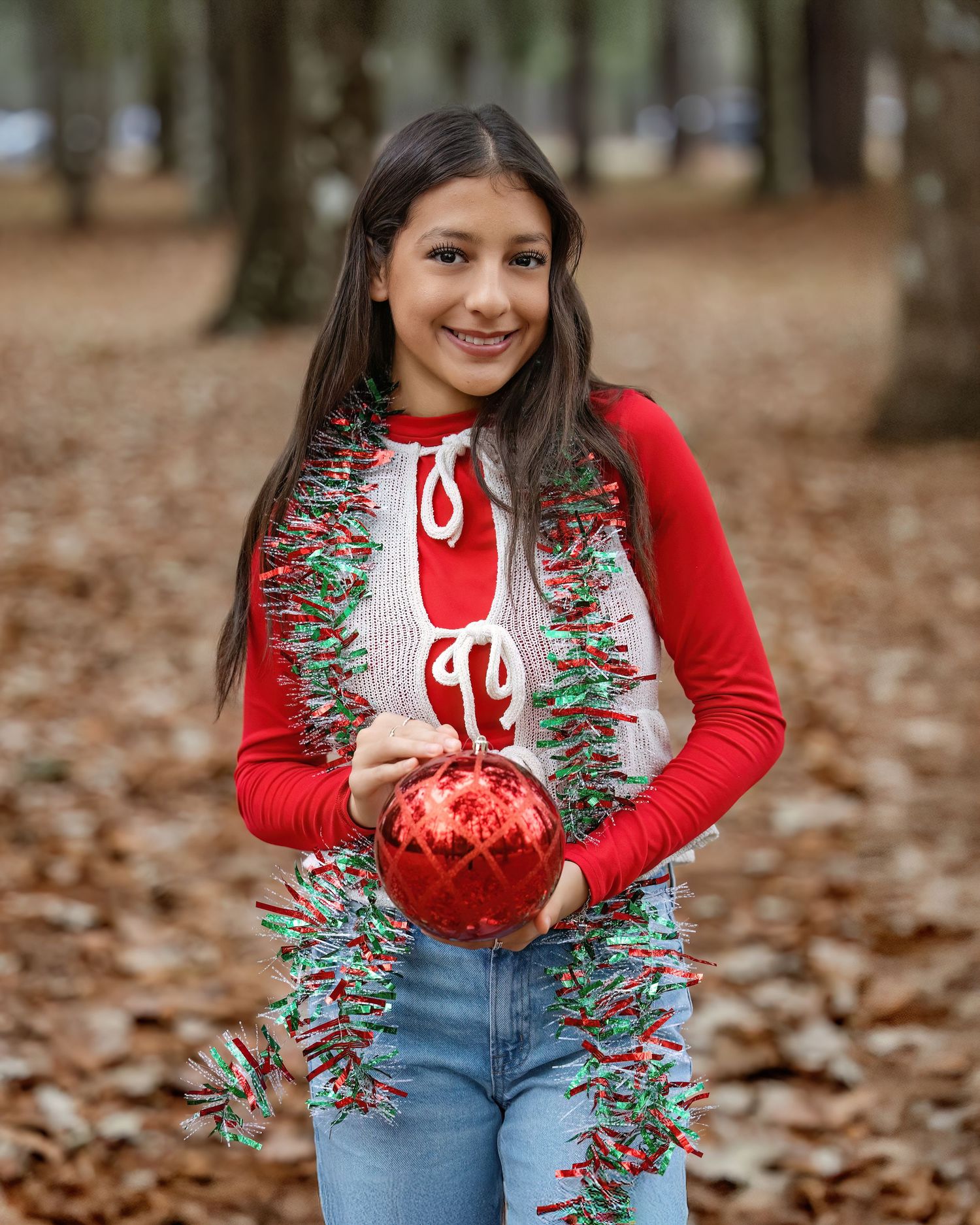 Marely BCHS Senior in a red Christmas sweater and jeans poses with festive tinsel garland in a forest of fallen autumn leaves.