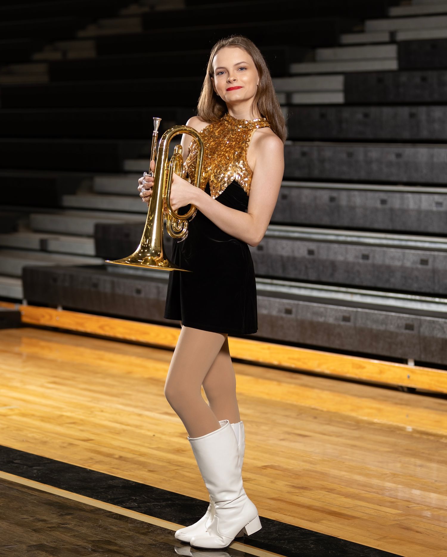 A performer in a black outfit and white boots holds a golden trophy while posing on wooden bleachers in a gym.