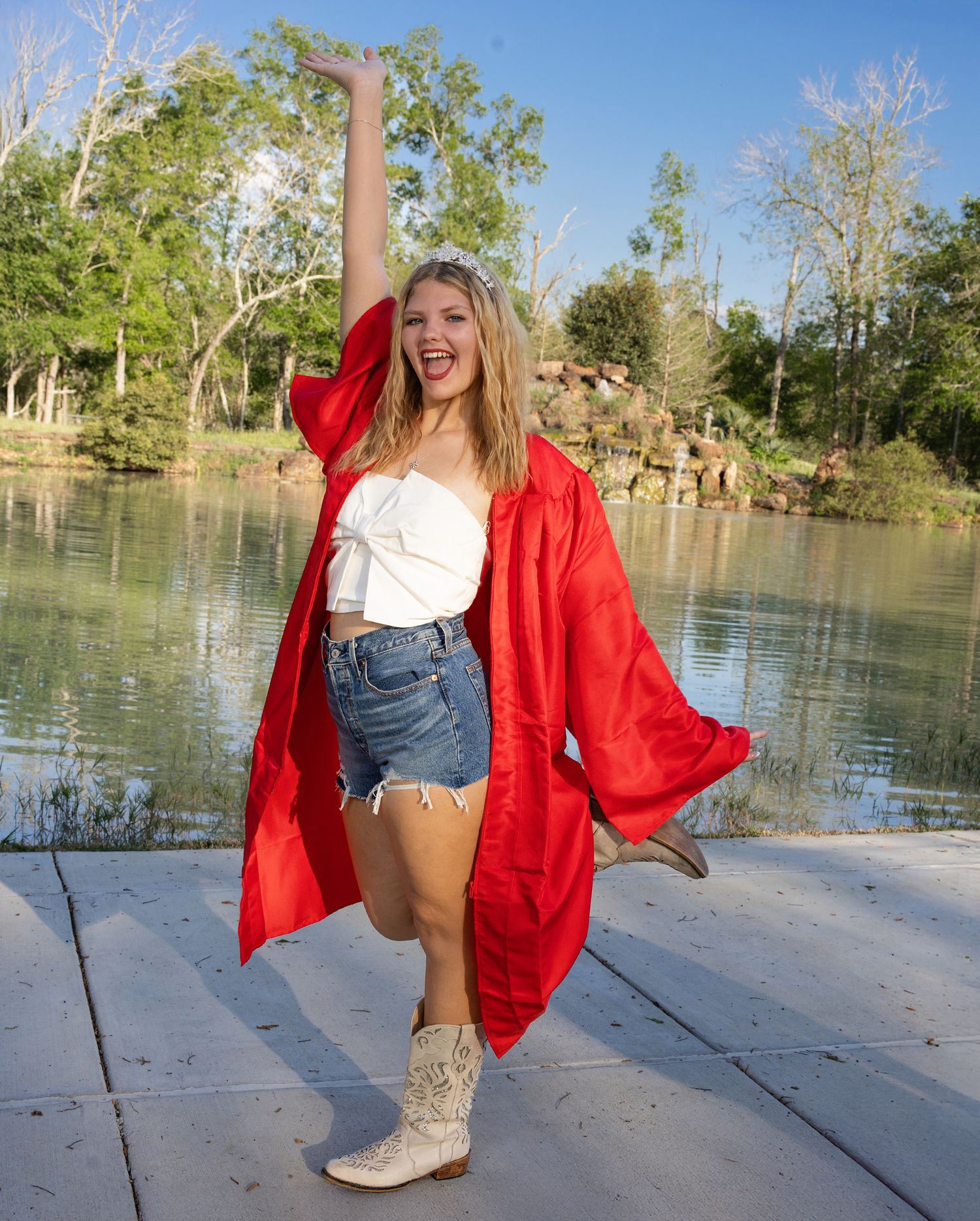 A figure in a bright red jacket and denim shorts poses playfully by a serene lake with bare trees in the background.
