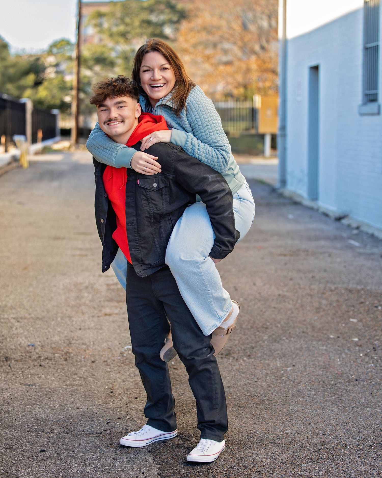 Two people share a playful piggyback moment outdoors on a sunny autumn day near a blue building.