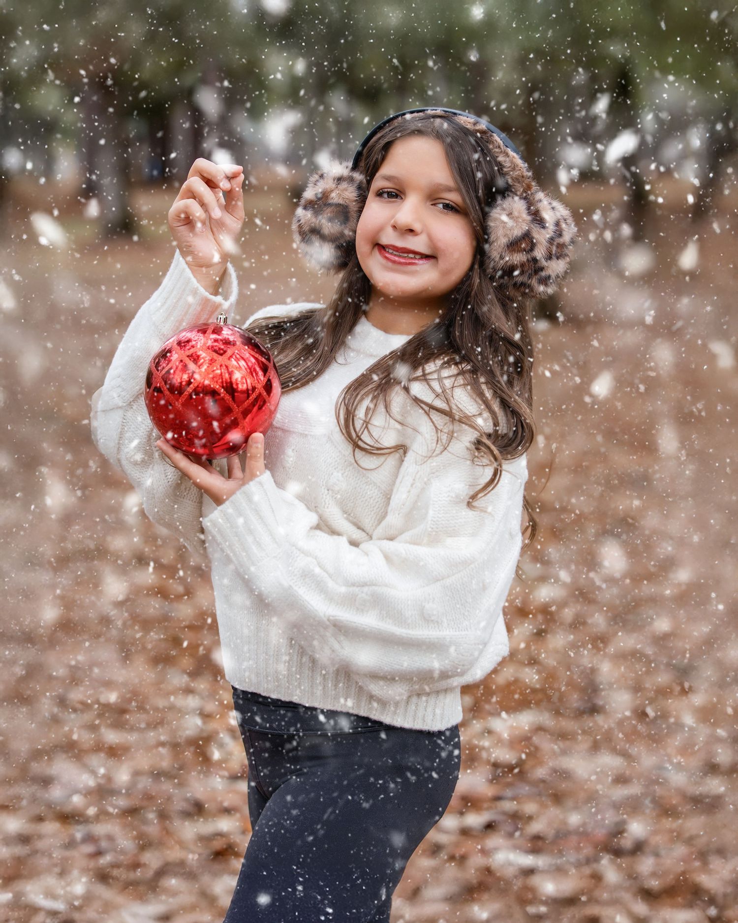 Child playing in falling snow wearing a white sweater and holding red Christmas ornaments.