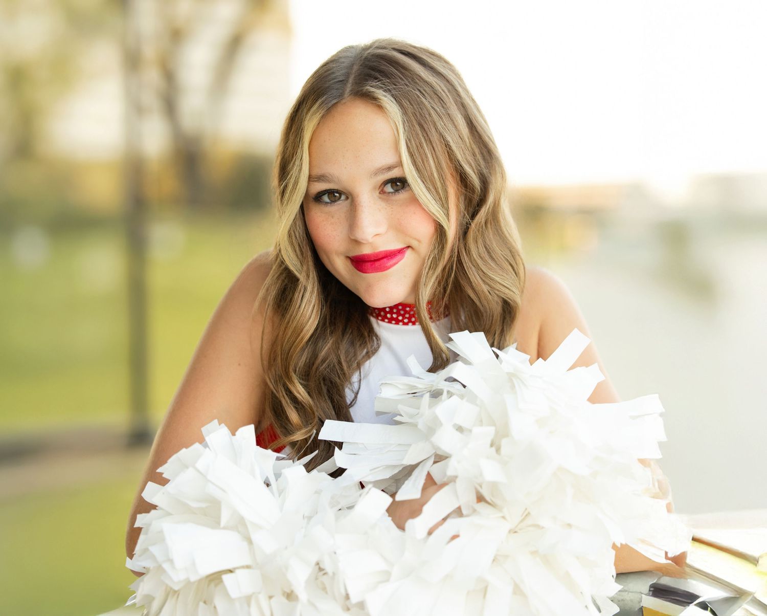 A cheerleader in a white uniform holds white pom poms while posing outdoors on a sunny day.